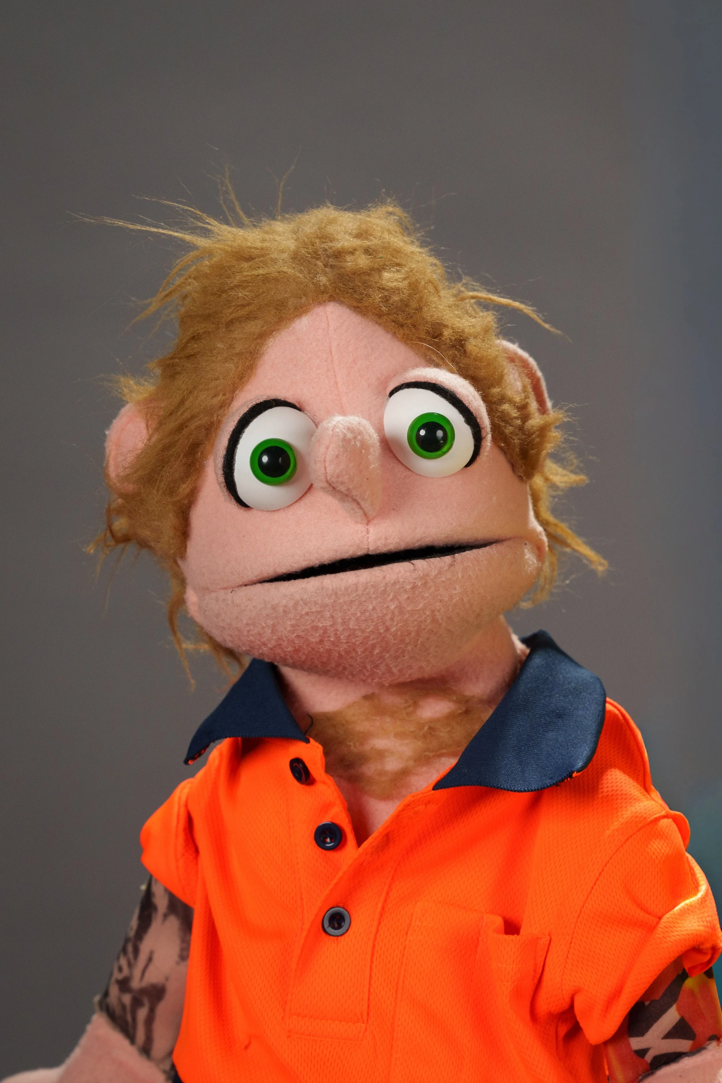 Close-up of puppet with brown curly hair, large green eyes, wearing an orange safety vest, against a plain grey background.