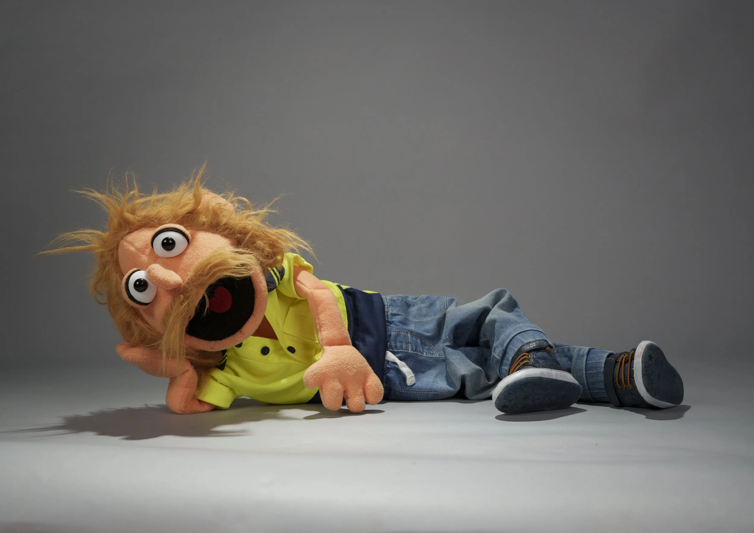 A puppet with blonde hair, large eyes, and a yellow shirt, lying on the ground.