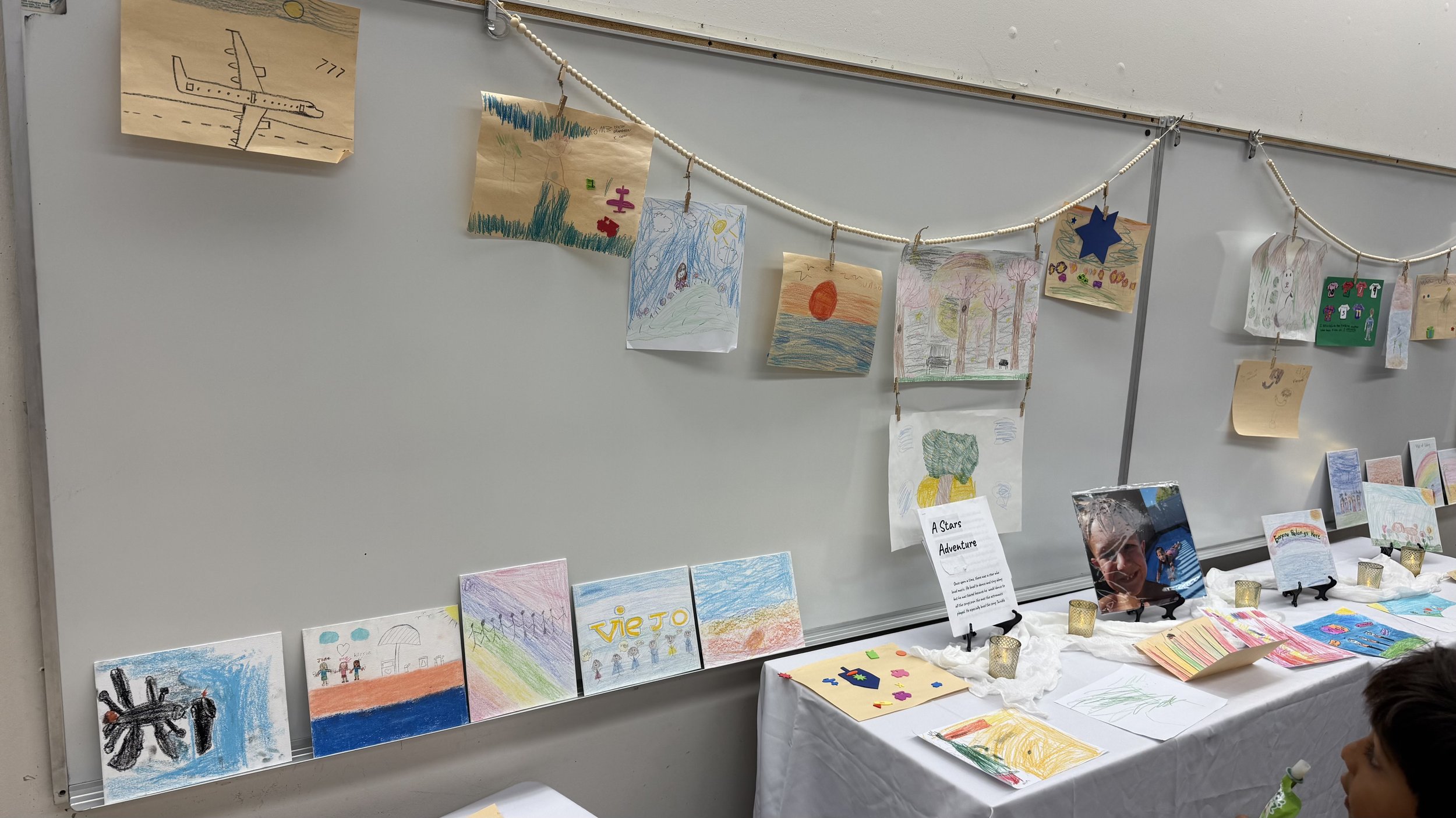 Children's artwork displayed on a whiteboard and table, featuring drawings of nature, animals, and colorful scenes, with some drawings hanging on a string with clothespins, and small decorative items on the table.