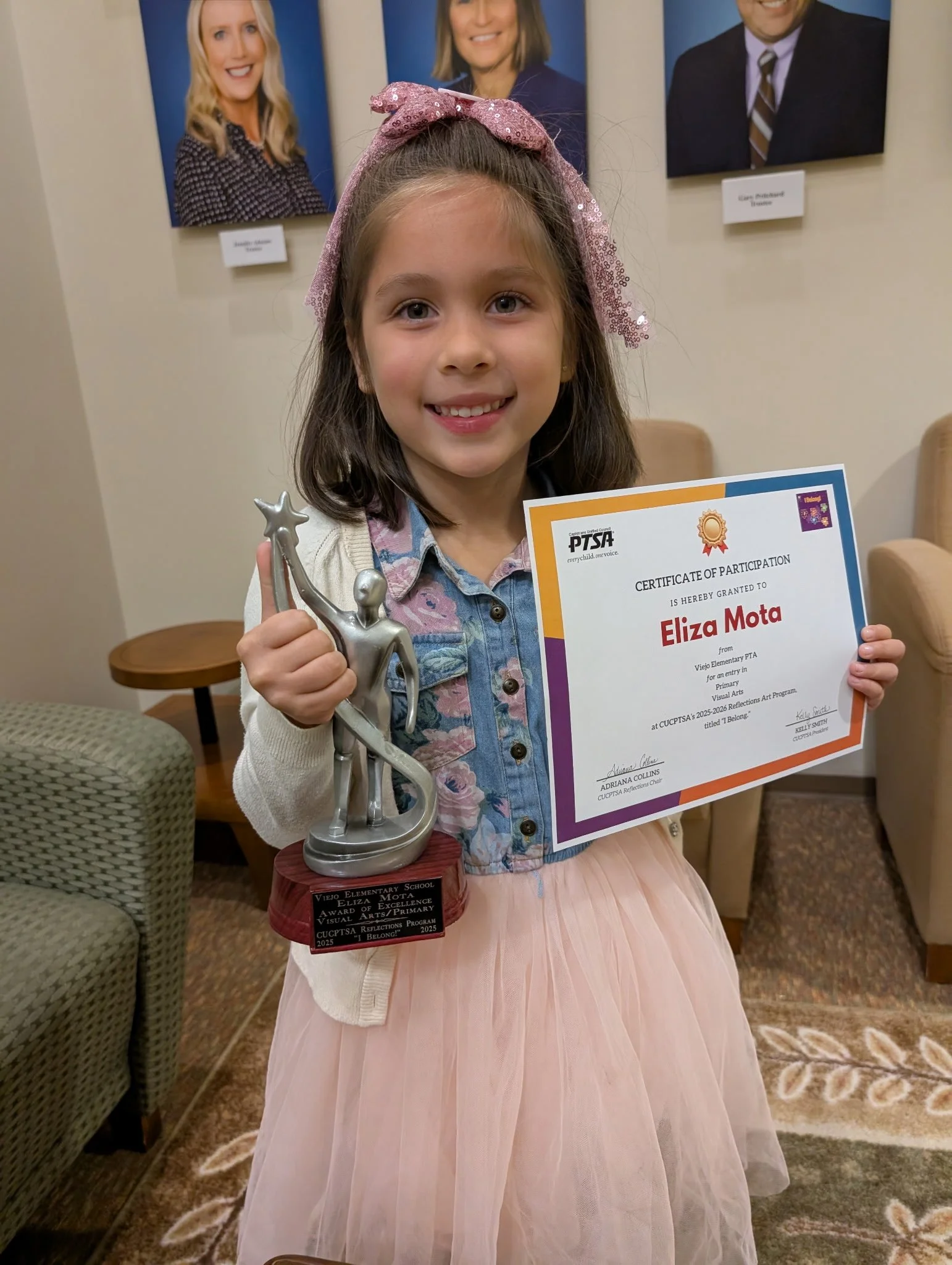 A young girl holding a trophy and a certificate, smiling at the camera, standing in a room with framed photos on the wall behind her.