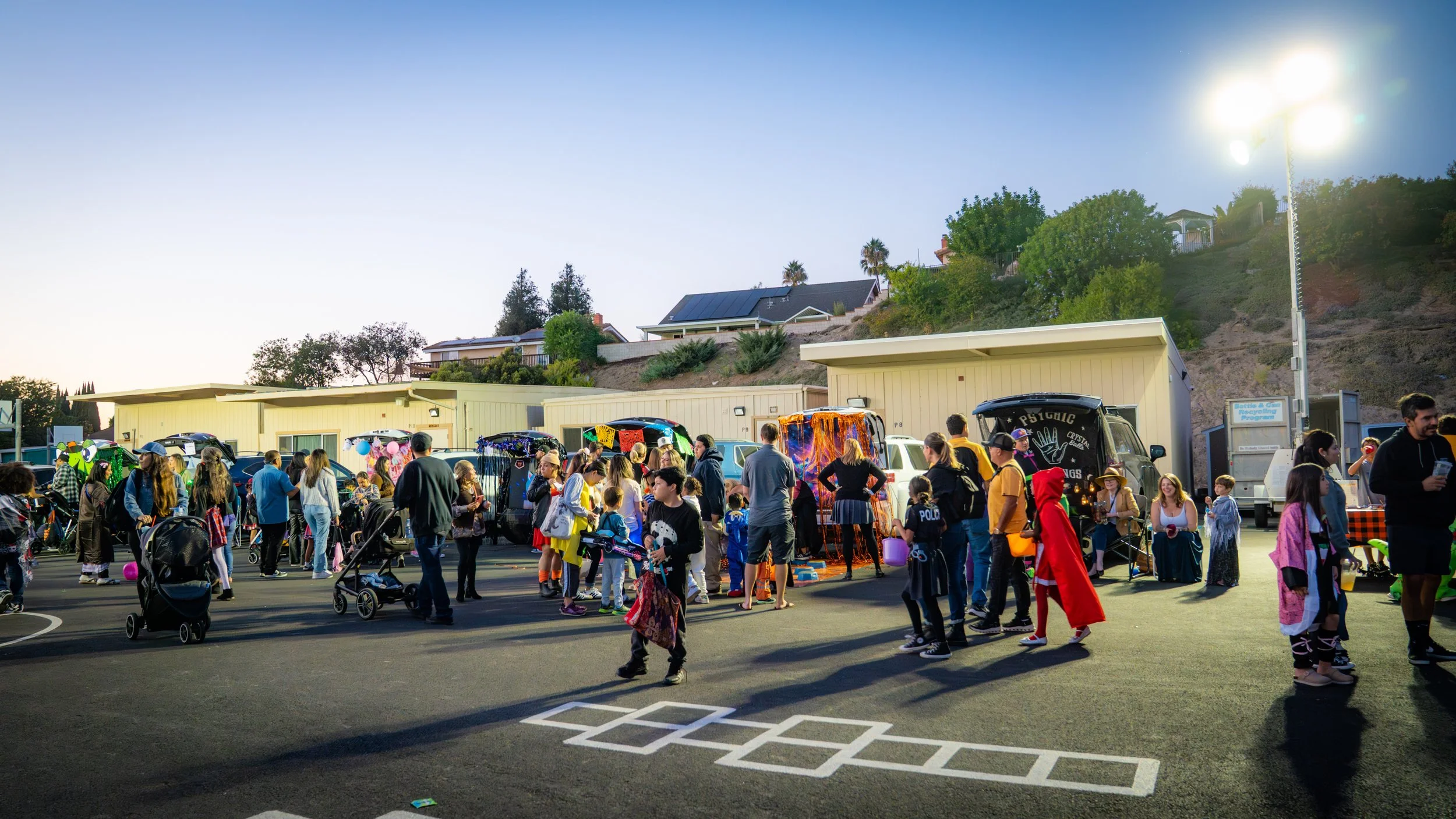 A festive outdoor event in a parking lot with people dressed in costumes, children with balloons, and decorated food trucks, evening setting with hill and houses in background.