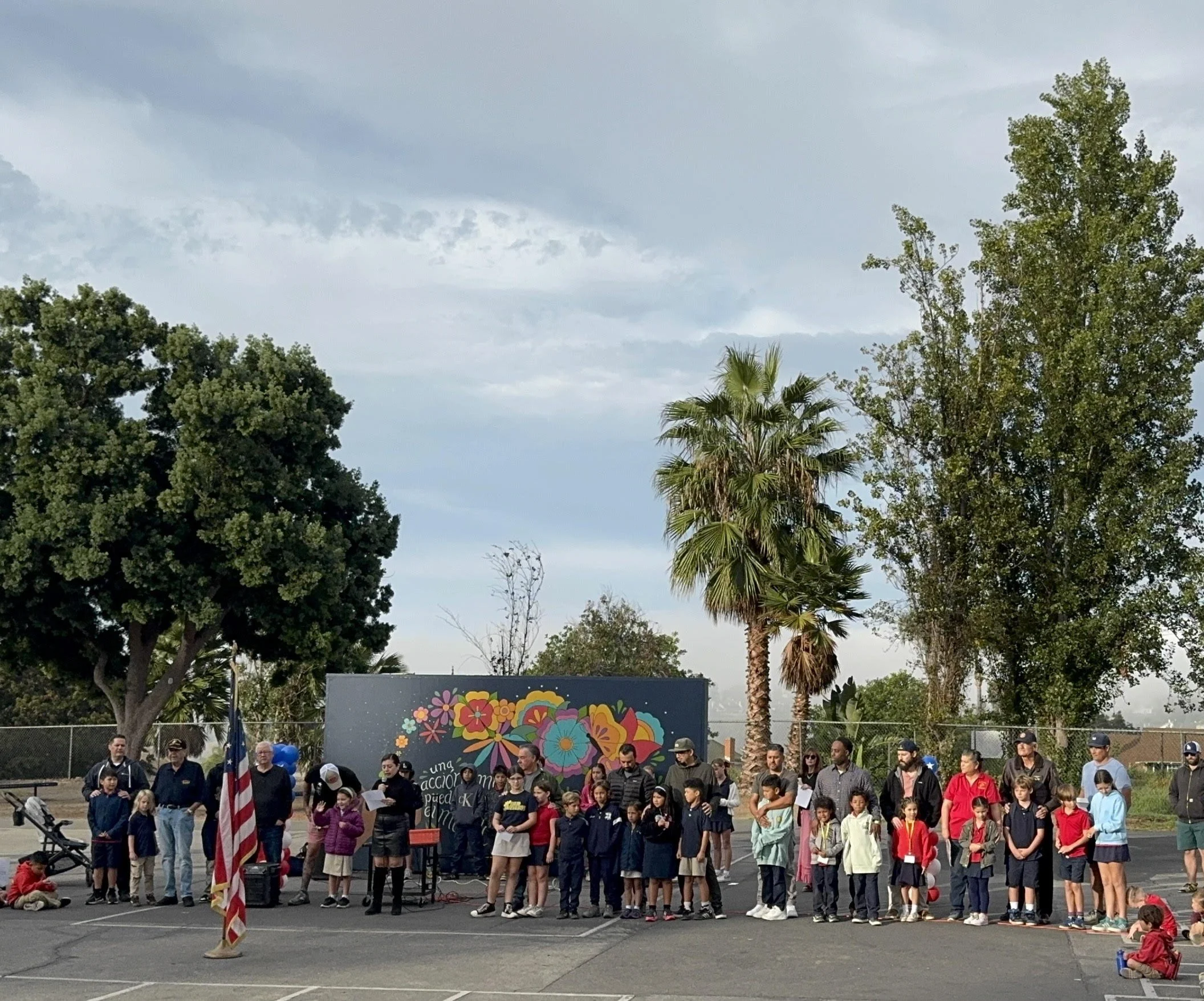A group of children and adults assembled outdoors near a black fence, with a colorful mural of flowers in the background, palm trees, and a cloudy sky overhead.