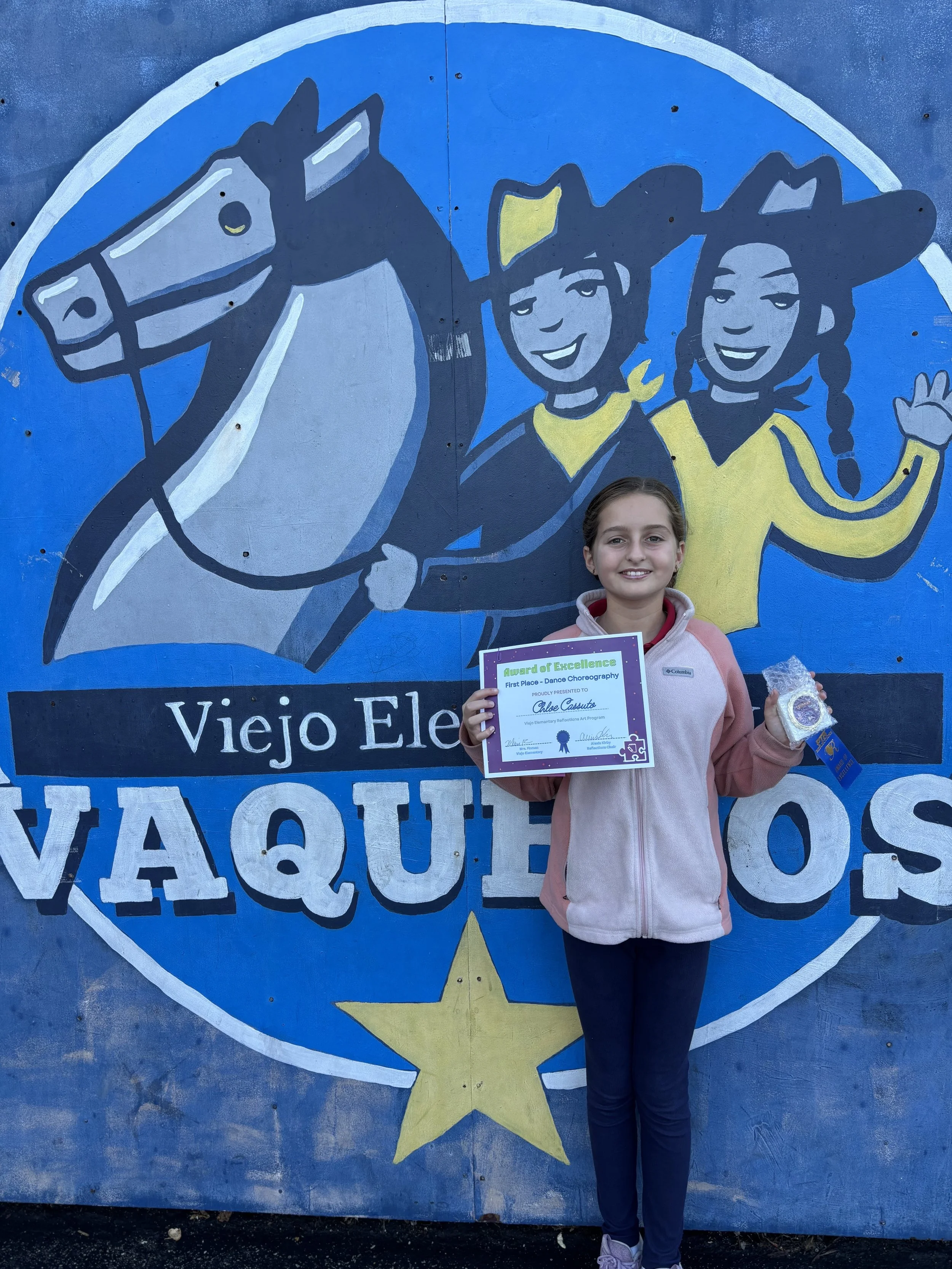A young girl standing in front of a colorful mural with a horse and two smiling children. She is holding a certificate and a medal, smiling at the camera.