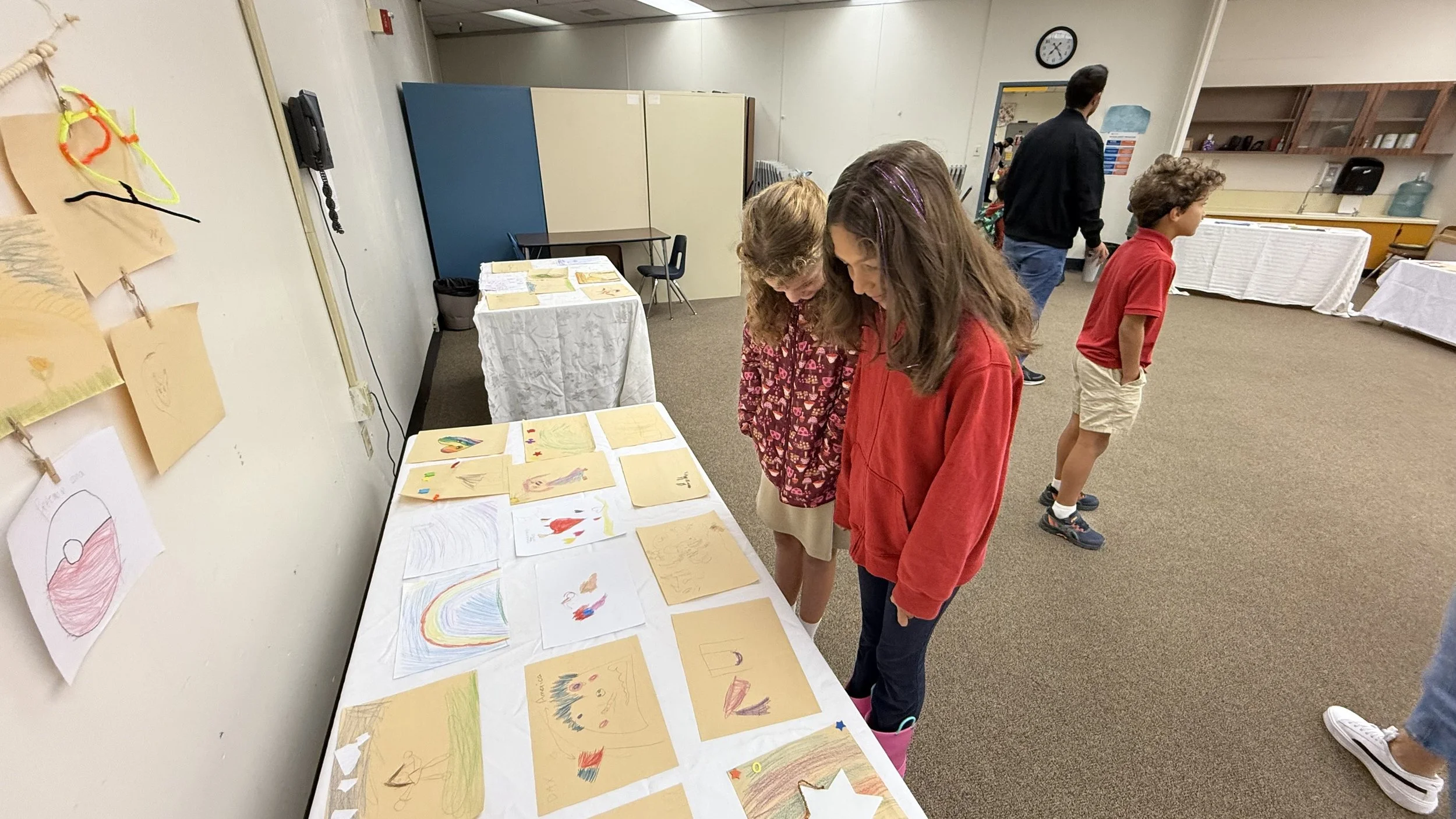 Three children are looking at drawings and artwork displayed on a table in a room, with other children and adults present in the background.