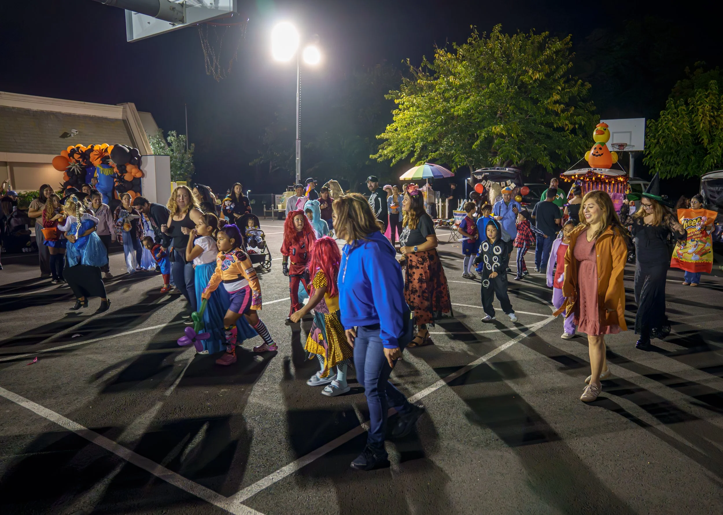 People, including children in costumes, dancing in a parking lot at night during a Halloween celebration, with Halloween decorations and a basketball hoop in the background.