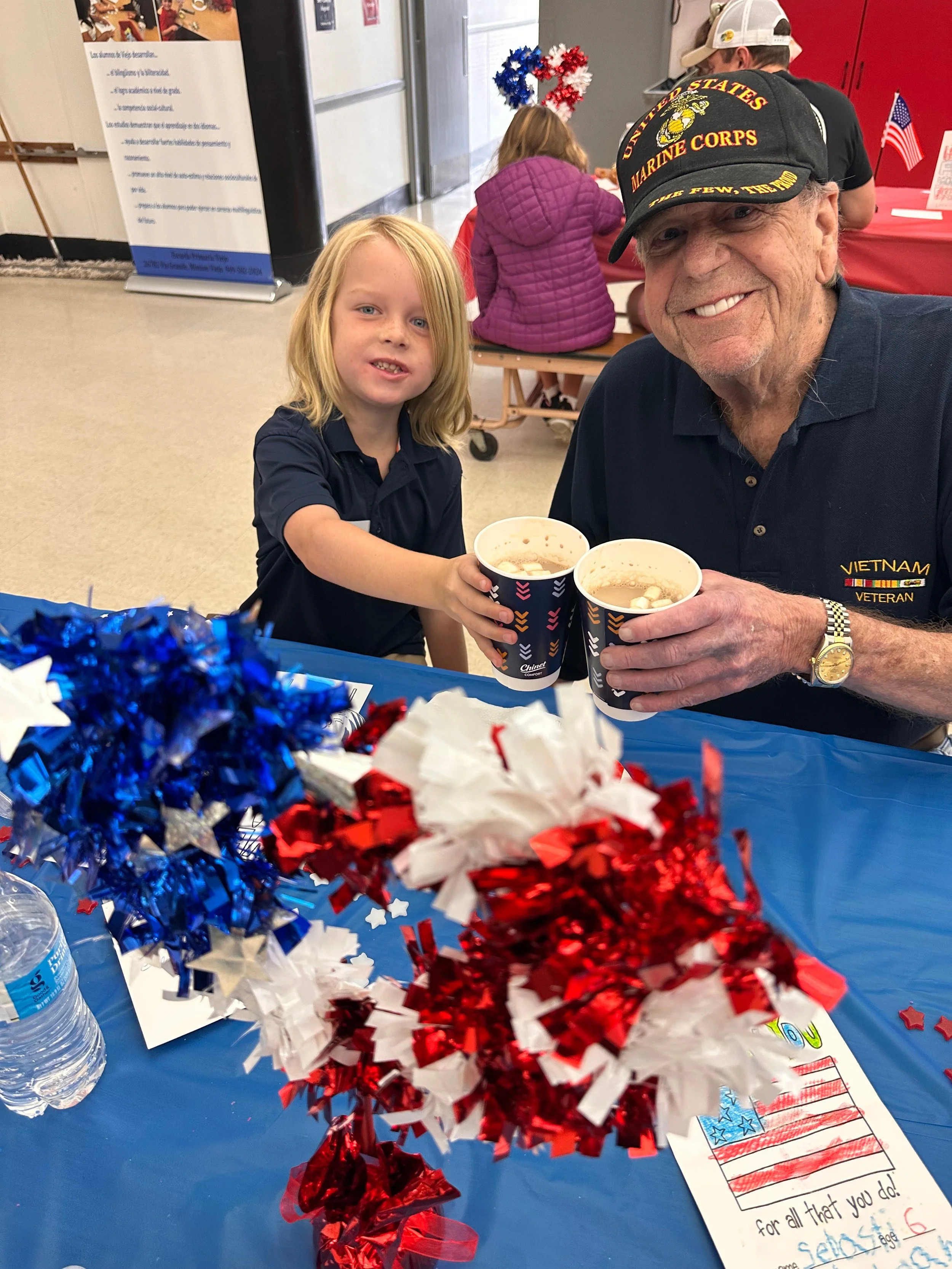 A young boy and an elderly man in military uniforms with patriotic decorations, holding cups of dessert and smiling at the camera, at a patriotic-themed celebration with red, white, and blue decorations and American flags.