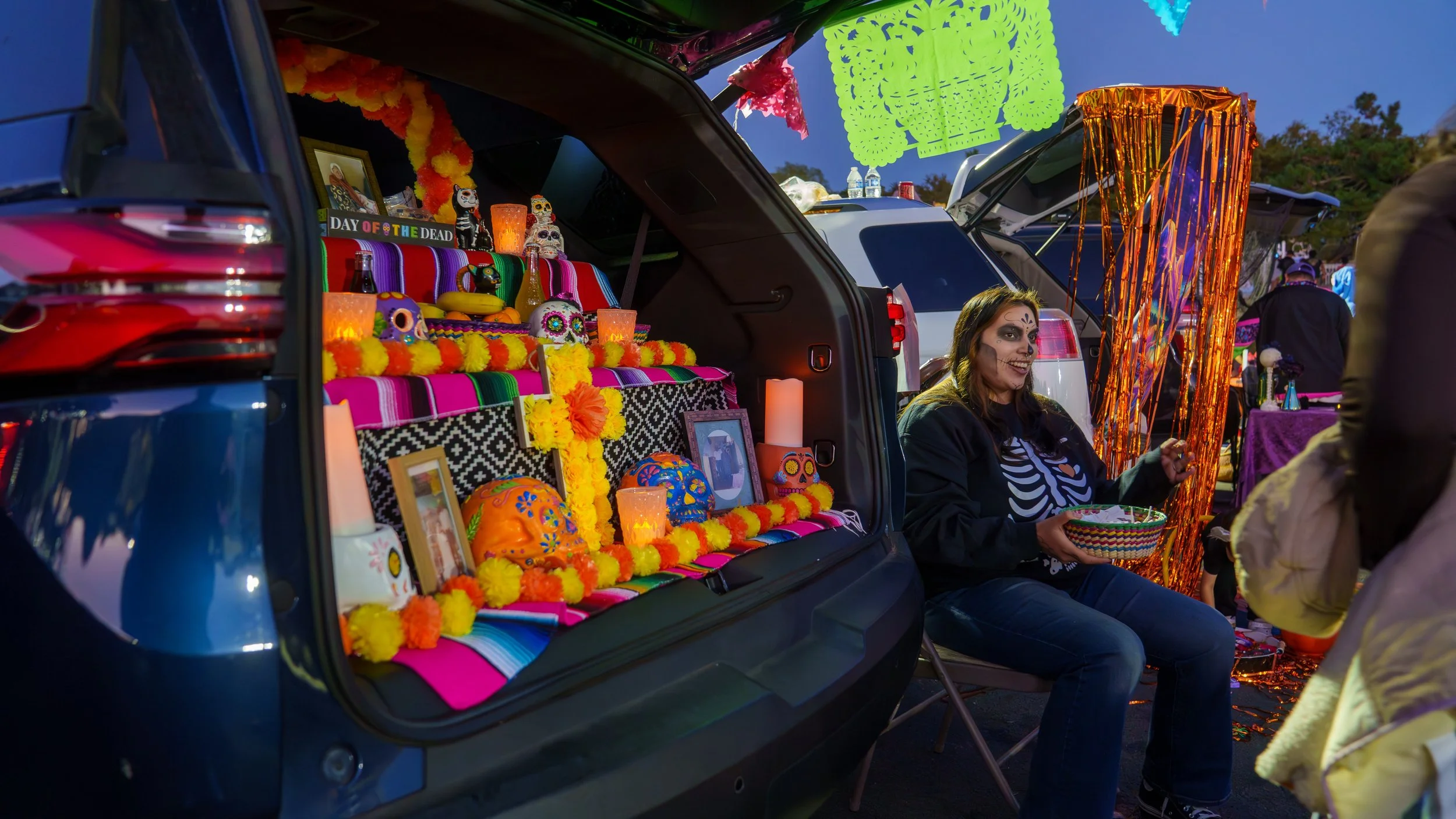 A woman dressed as a skeleton with face paint sits outside near a decorated car trunk and a colorful carnival-style booth, celebrating Día de los Muertos with flowers, candles, and Día de los Muertos symbols in the evening.