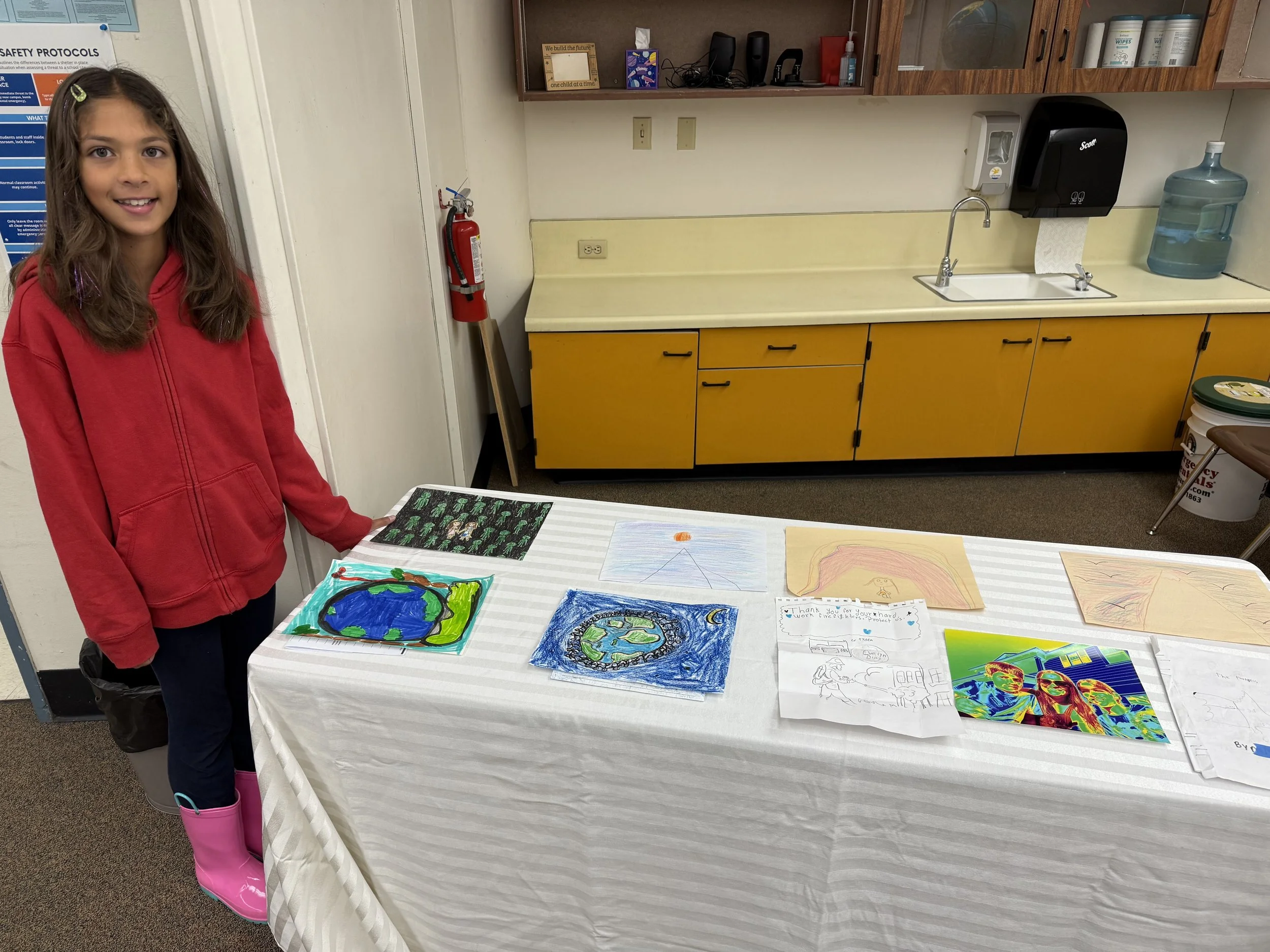 A young girl standing beside a table displaying various colorful drawings on paper, in a room with yellow cabinets, a sink, and cleaning supplies.