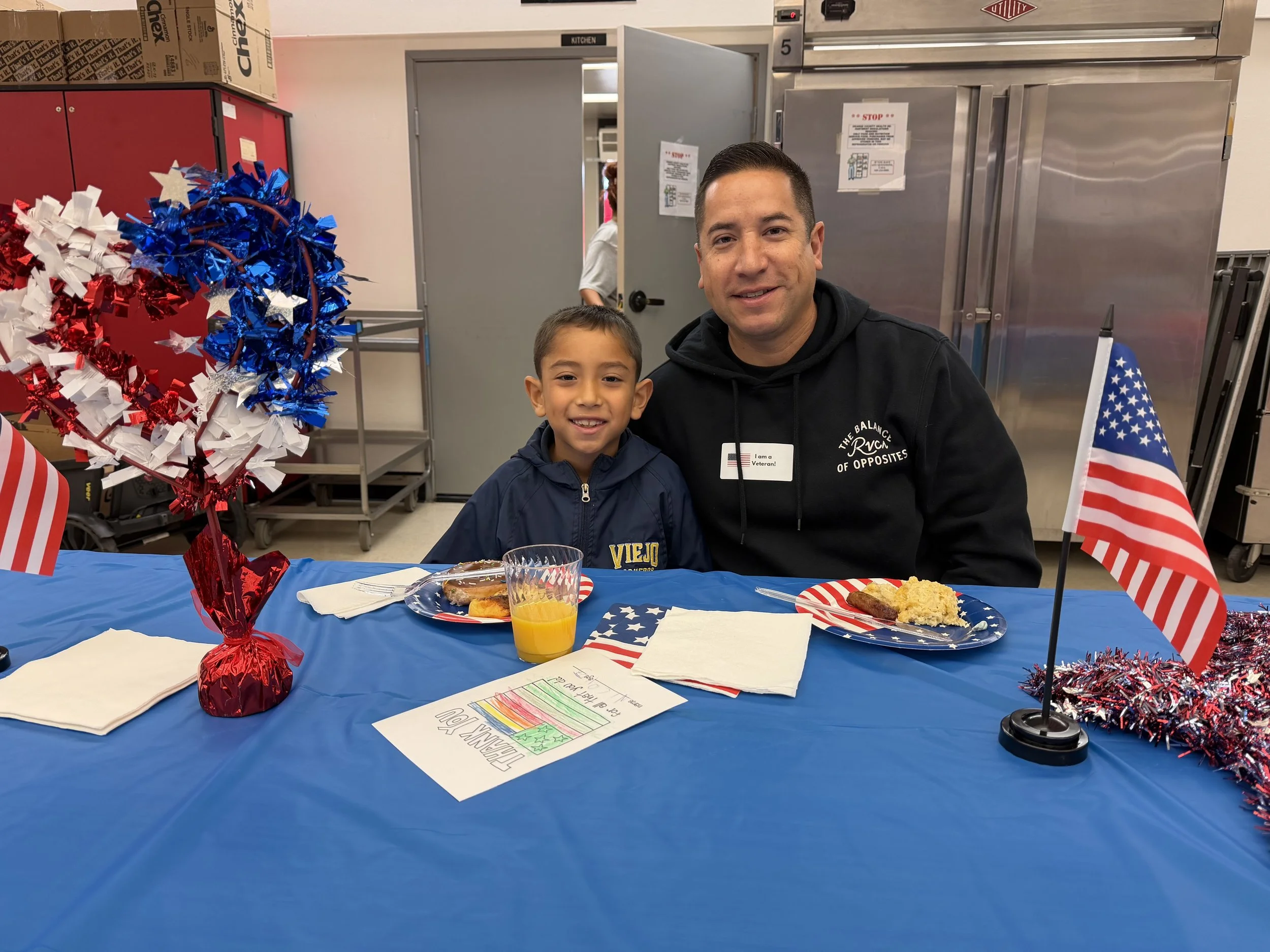 A man and a young boy sitting at a table decorated with American flags and patriotic decorations, celebrating a holiday, with plates of food and a glass of orange juice in front of them.