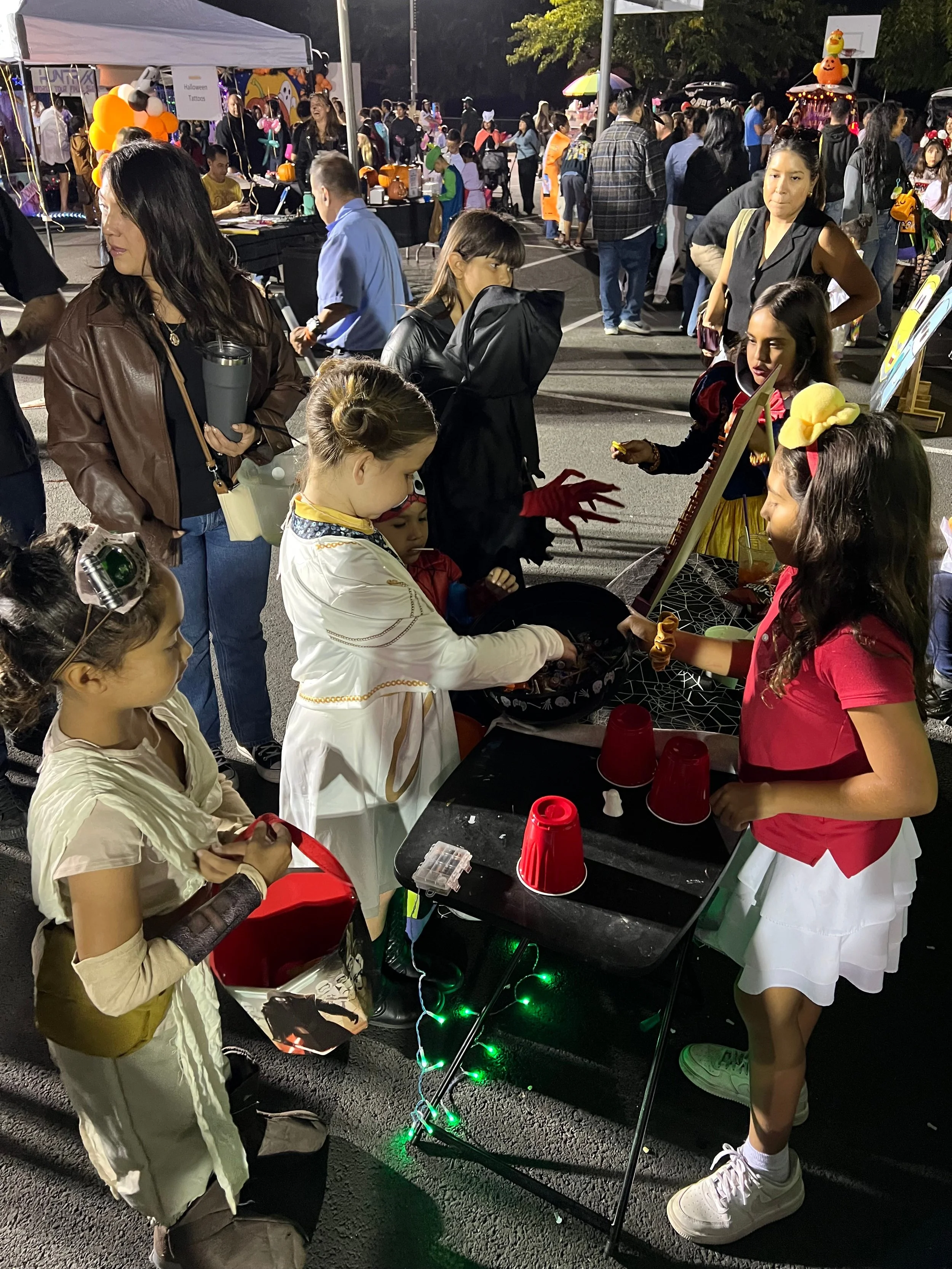 Children and adults dressed in Halloween costumes at a nighttime outdoor event with booths and decorations, including pumpkins and scarecrows.