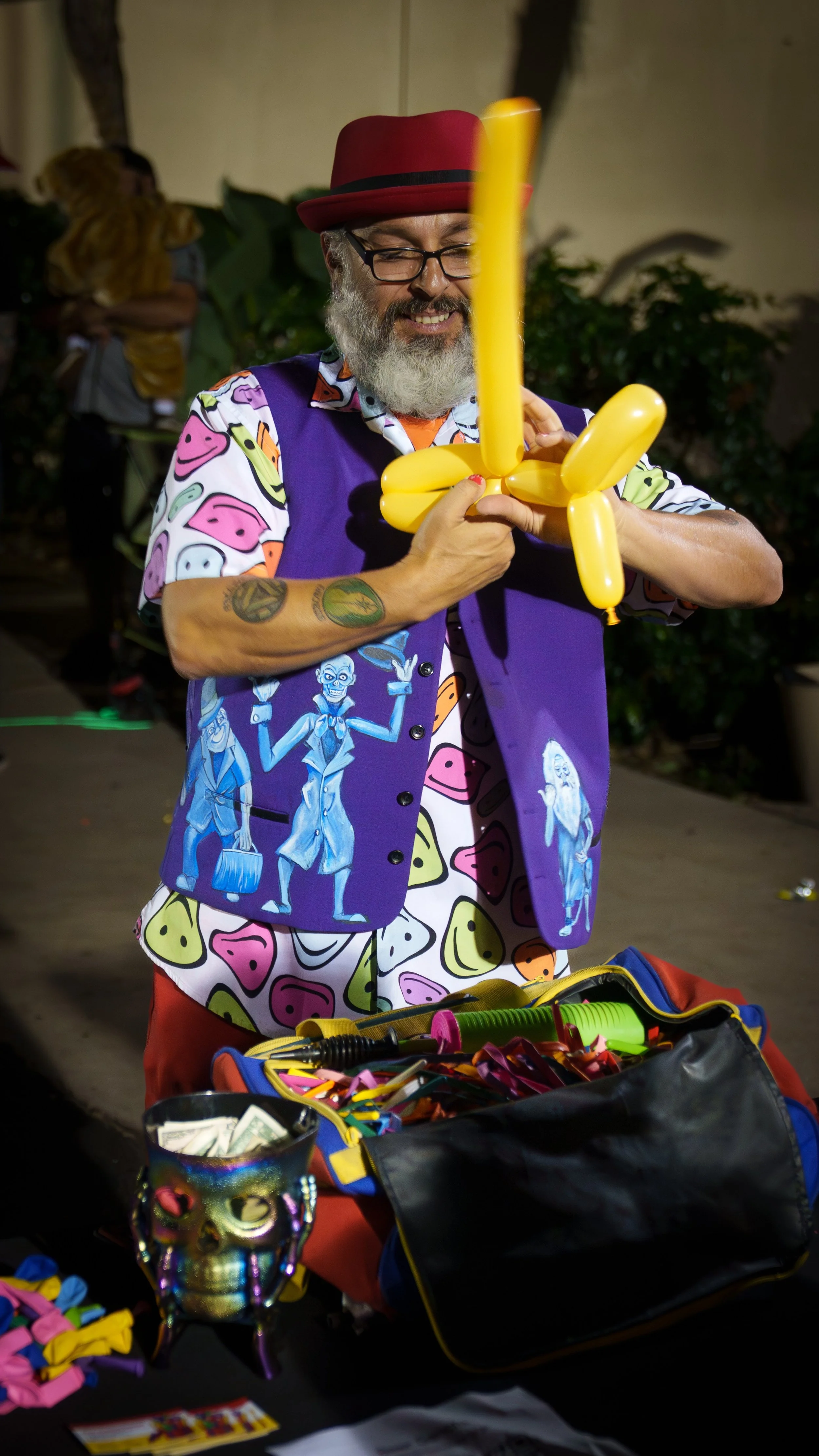 Man with a beard and glasses wearing a colorful shirt with smiley faces, a purple vest with cartoon skeletons, and a red hat, making balloon animals at a party or event.