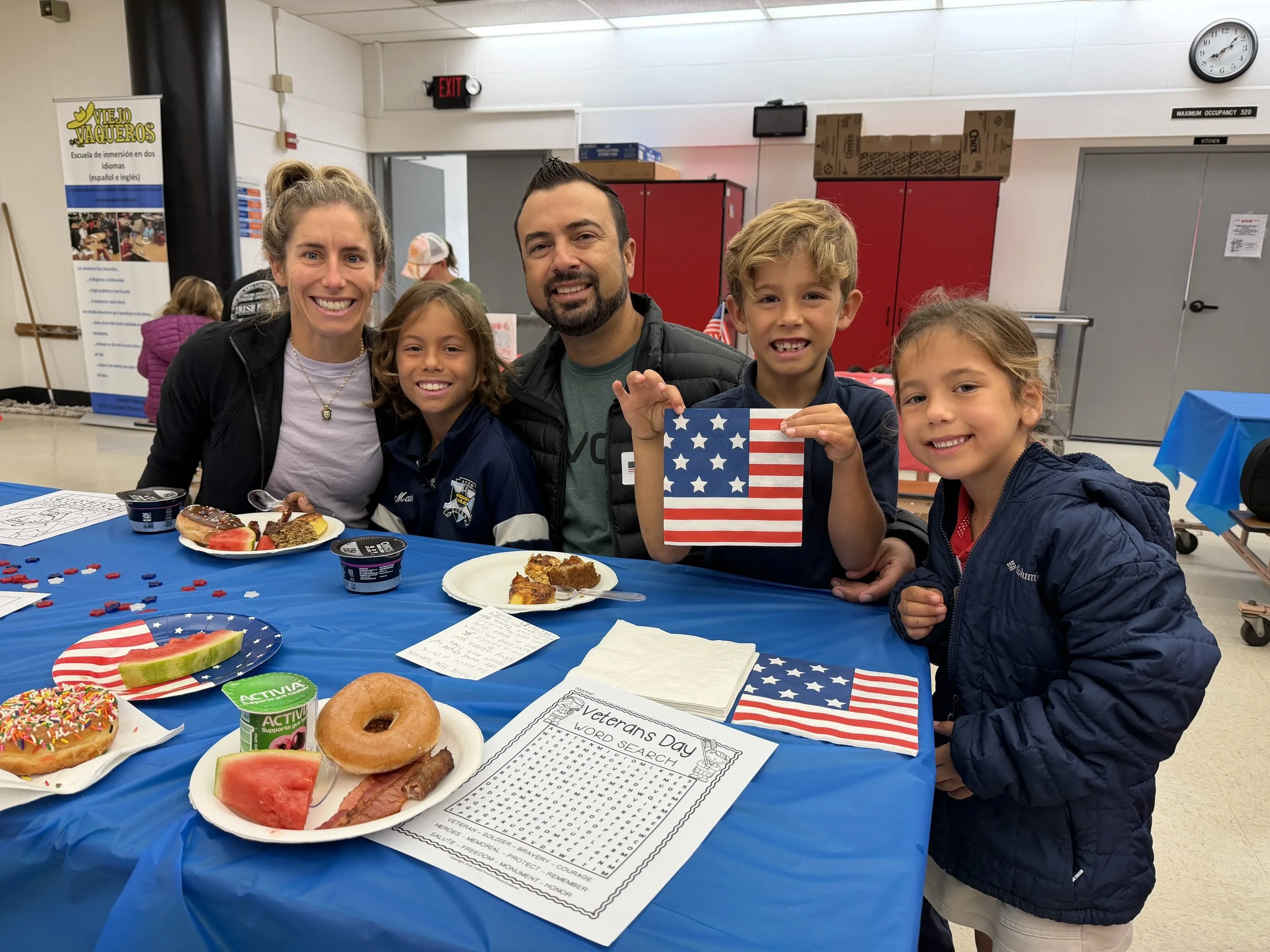 A group of five people, two adults and three children, sitting at a table decorated for Veterans Day, smiling for the camera. The table has American flags, donuts, watermelon, and a worksheet with a word search about veterans.