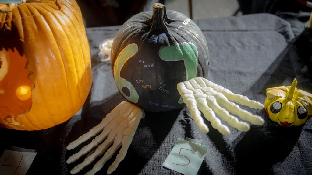 Decorative pumpkins, including one painted with numbers and signals, and skeleton hand and face decorations, on a dark fabric surface.