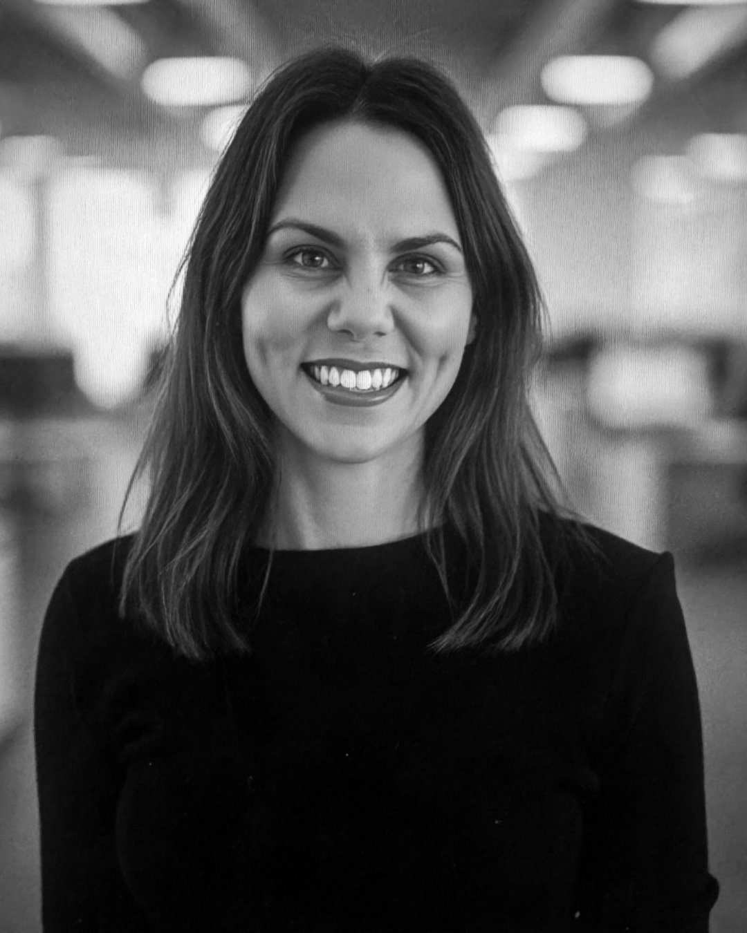 Black and white photo of a smiling woman with medium-length hair, wearing a dark top, standing in an indoor setting with blurred lights in the background.