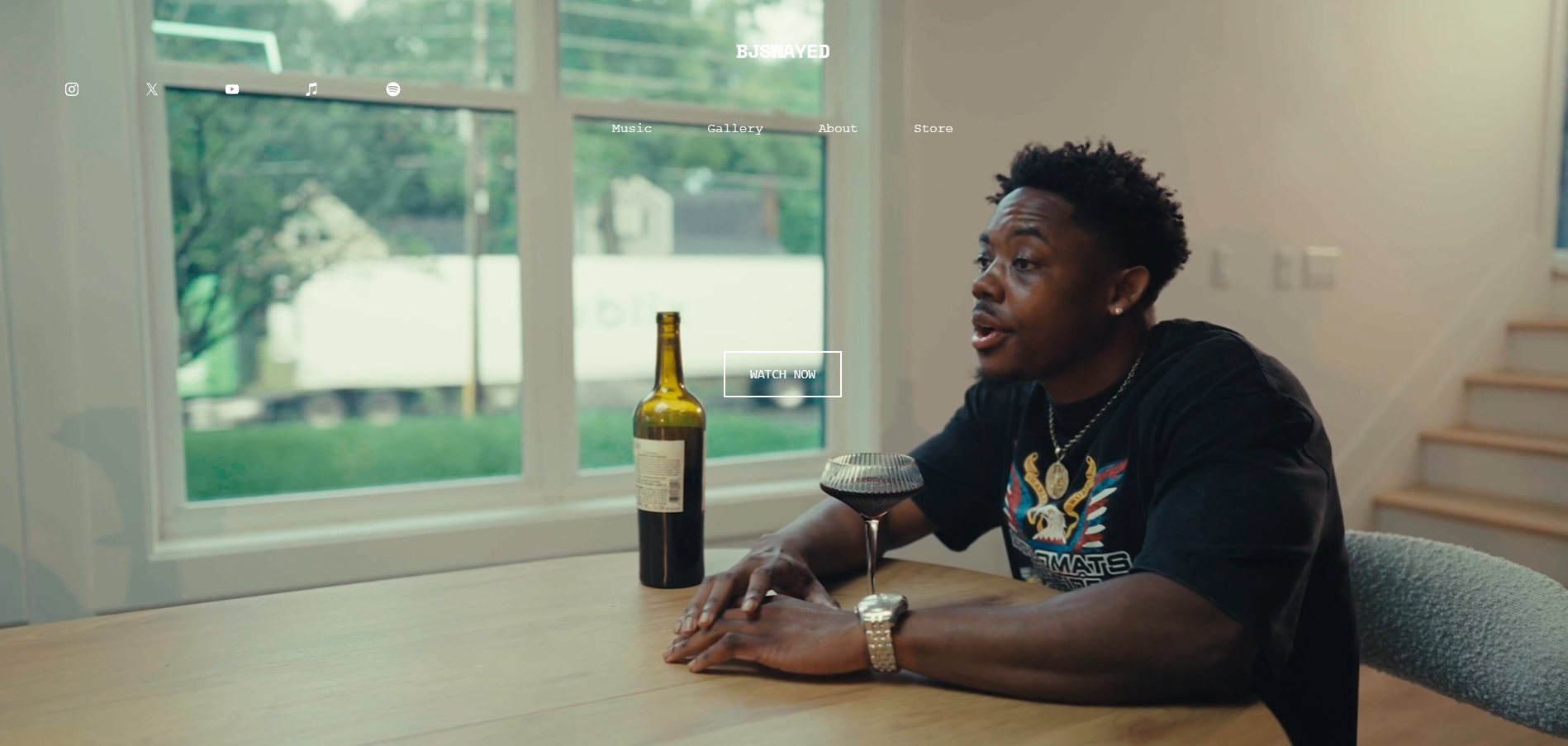 A man sitting at a dining table with a bottle and glass of red wine, inside a house with big windows showing greenery outside.