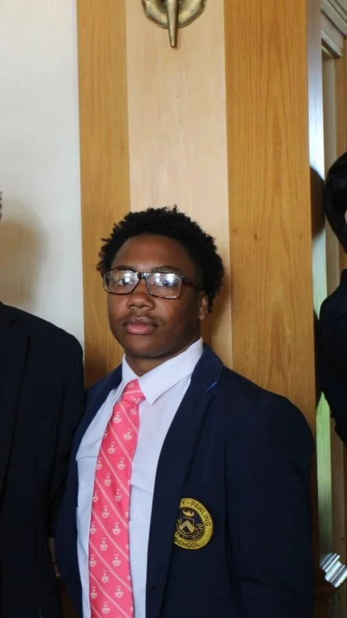 Young man wearing glasses, a white shirt, pink patterned tie, and navy blazer with school emblem, standing indoors against a wooden wall.