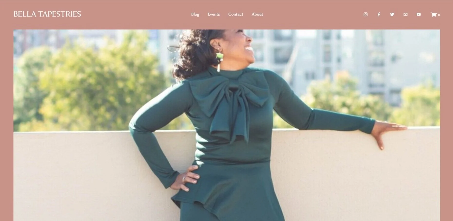 A woman wearing a dark green dress with a bow on the shoulder, smiling and leaning on a white balcony railing outdoors with a blurred background of trees and buildings.