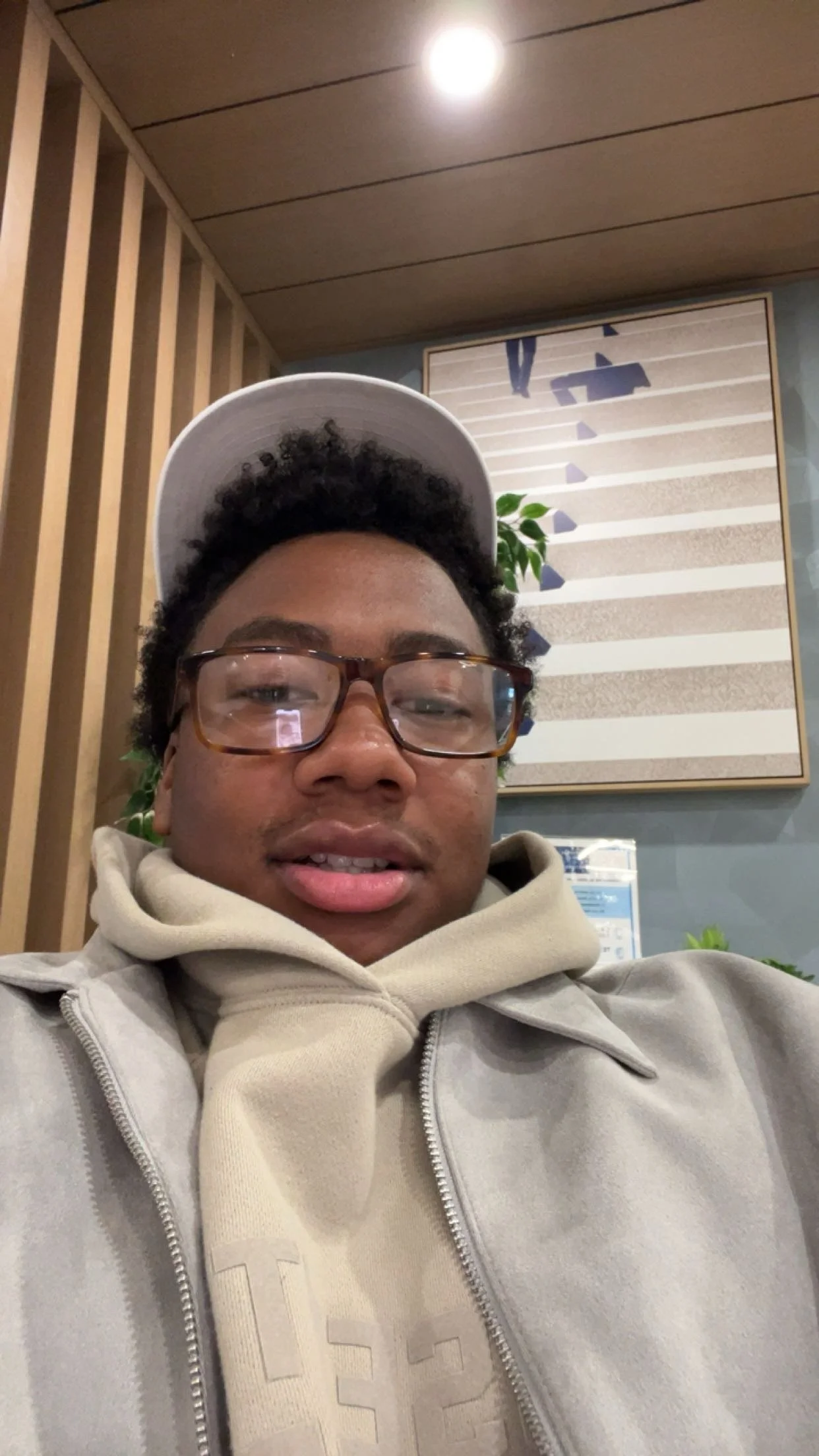 Young man wearing glasses, hoodie, and a white cap taking a selfie indoors.