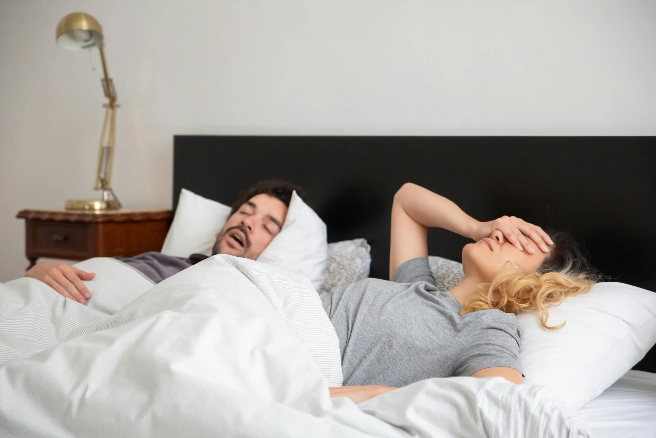 Woman in bed holding a hand against her face next to a snoring man