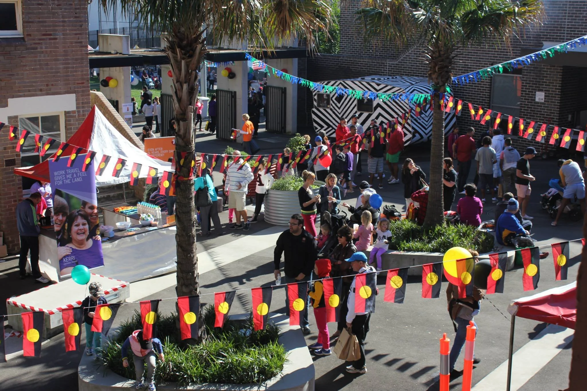 Outdoor event with people gathered around booths and decorations, including banners, balloons, and flags, in a courtyard area with trees and brick buildings.