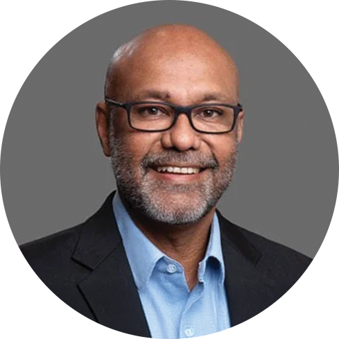 A middle-aged man with glasses, a beard, and a bald head, smiling, wearing a dark blazer and a light blue dress shirt against a gray background.