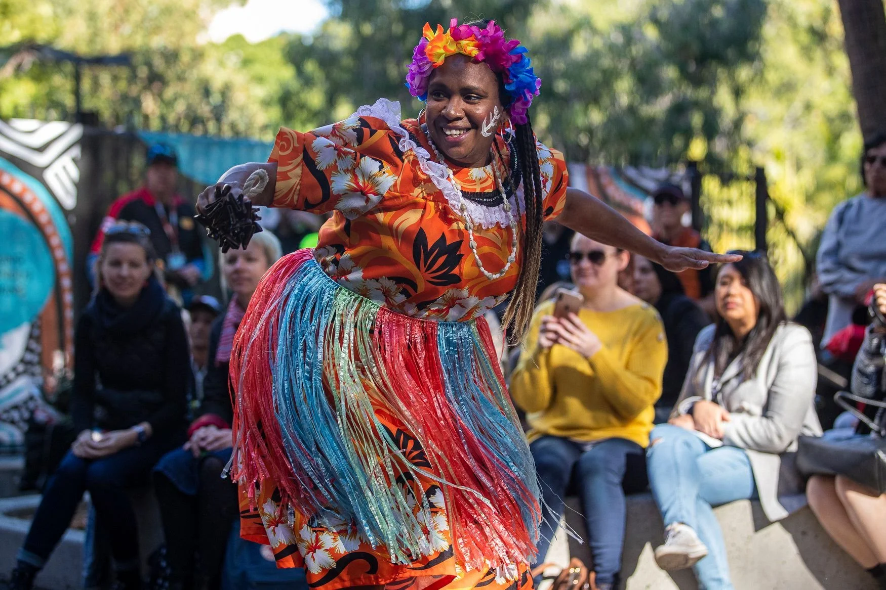 A woman in colorful, tropical-themed clothing and a rainbow flower crown performs a dance outdoors, with a crowd of onlookers seated around her.