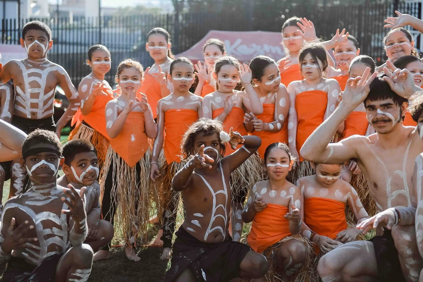 Group of children and a man with traditional face paint, dressed in costumes made of natural materials, gathered outdoors for a cultural event or performance.
