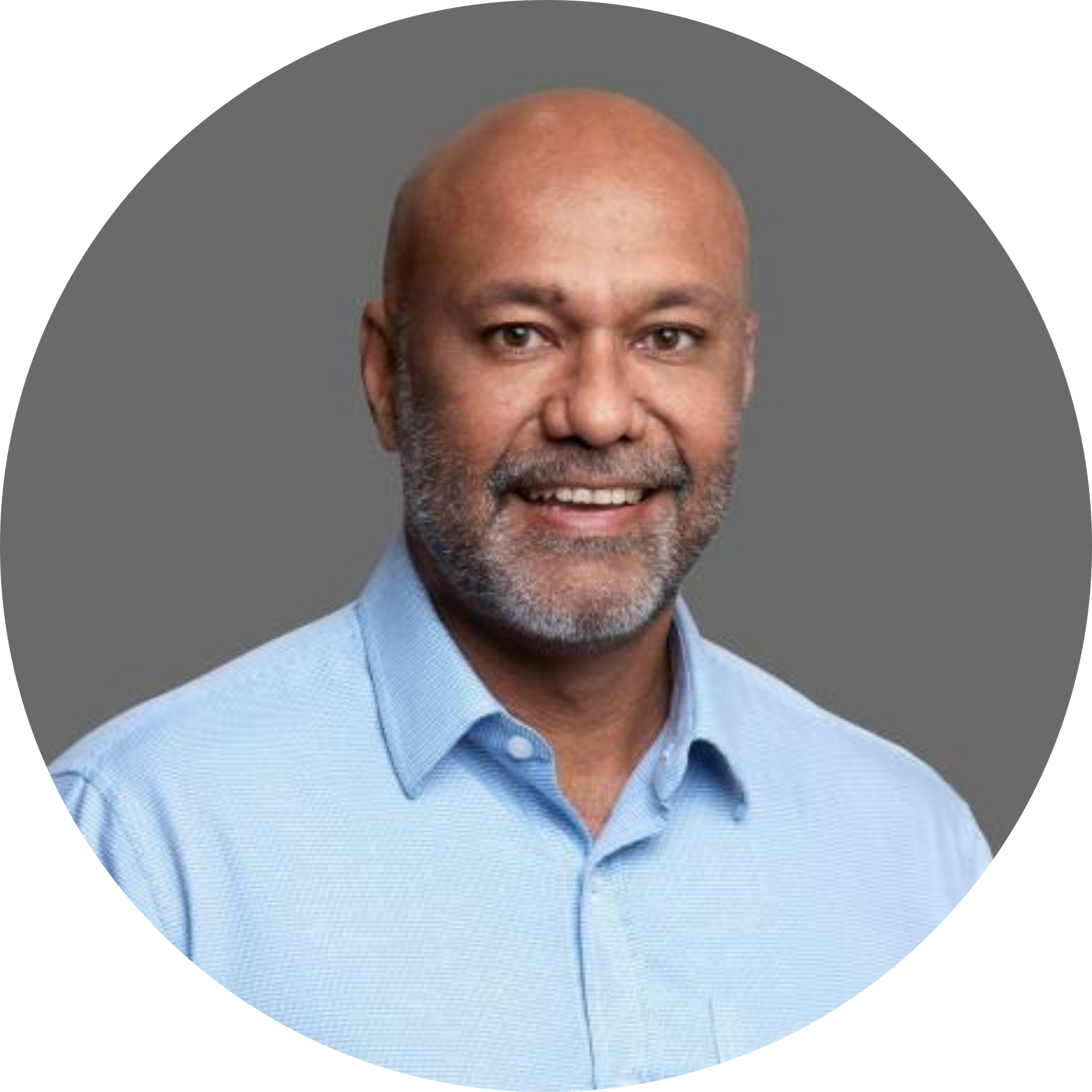 Circular headshot of a bald man with a gray beard, smiling at the camera. He's wearing a light blue button-down shirt against a plain gray background.