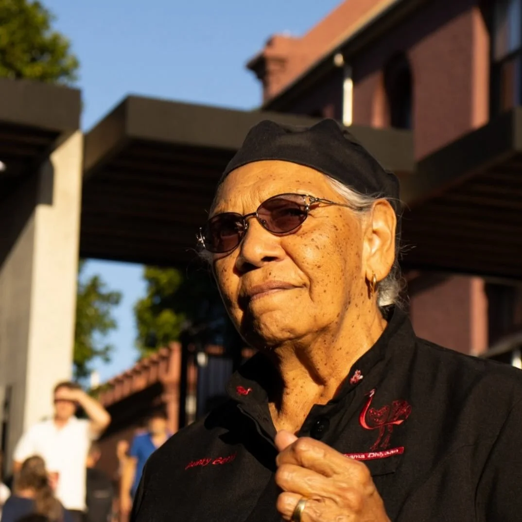 An older woman with short gray hair, wearing sunglasses, earrings, a black hat, and a black shirt with red embroidery, stands outdoors on a sunny day, holding her collar.