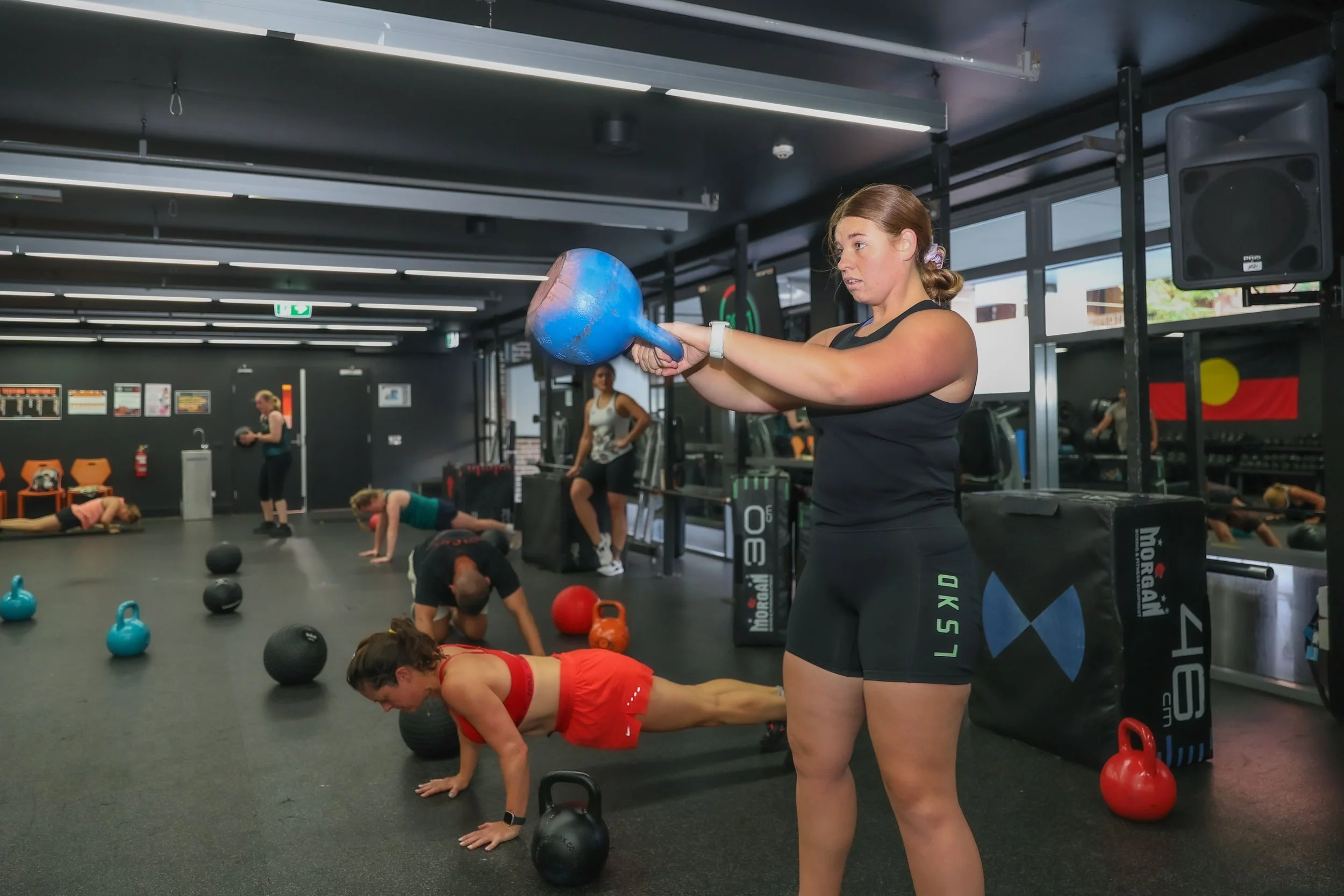 A woman in black workout clothes lifting a blue kettlebell in a gym, with multiple people in the background performing various exercises involving kettlebells and bodyweight exercises.