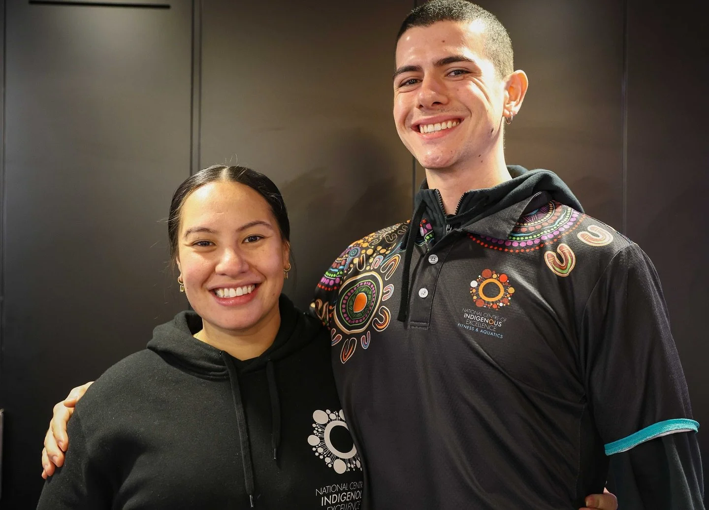 Two smiling young people, a woman and a man, standing close together with their arms around each other, wearing black hoodies and athletic shirts with colorful designs, in front of a dark background.