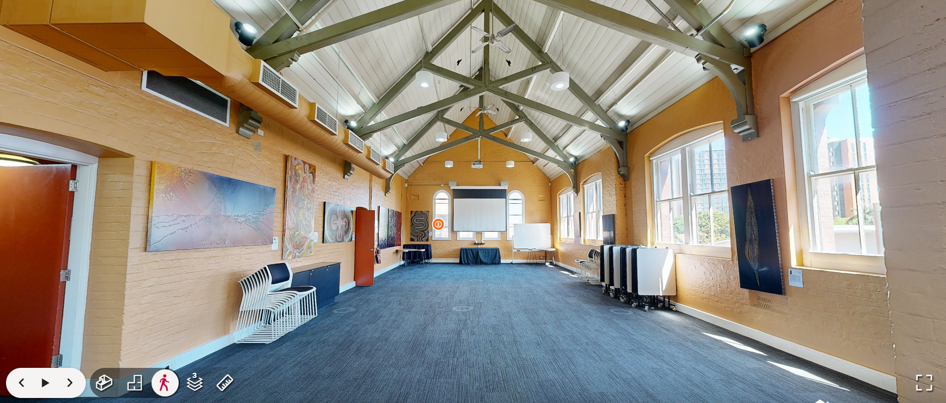 Empty art gallery with colorful paintings on yellow brick walls, large windows, and a white projection screen at the far end, with chairs stacked on the right side and a table near the front.