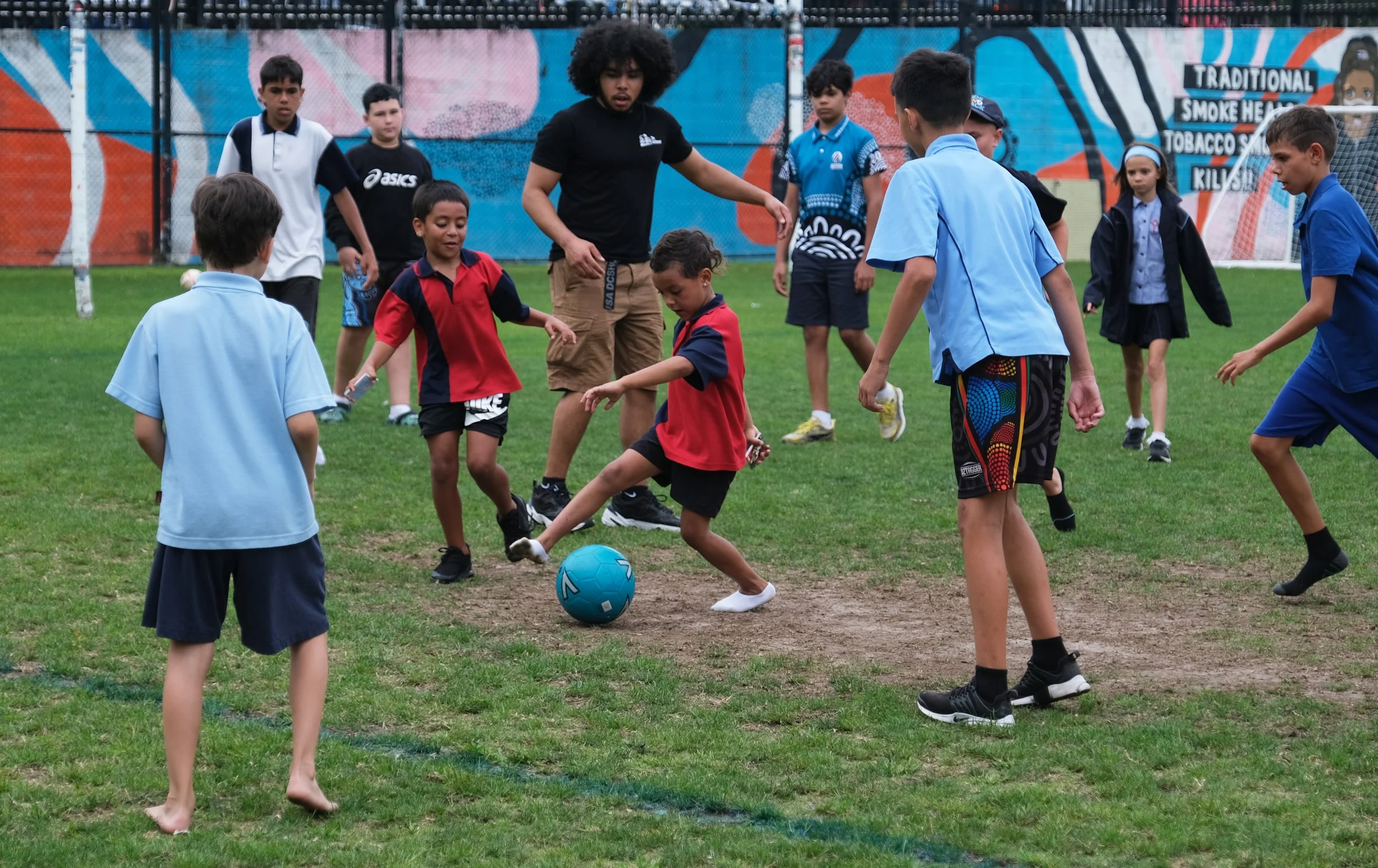 Group of children playing soccer on a grassy field with a fence and colorful mural in the background.