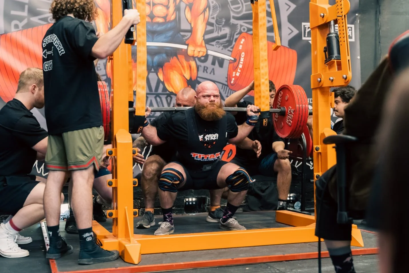 A man with a beard performing a squat with a heavily loaded barbell at a powerlifting competition, surrounded by spotters and spectators in a gym setting.