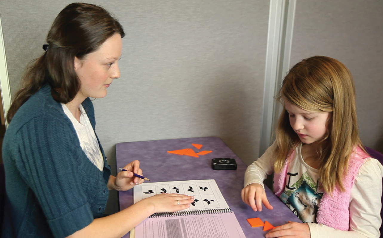 A woman and a girl sit at a table playing a math game with shapes and tiles, with a worksheet and a camera on the table.