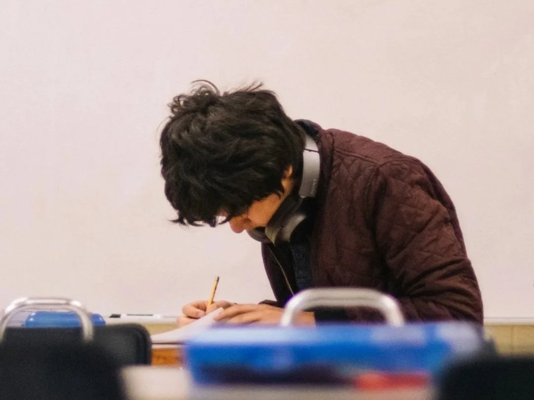 Person with curly hair wearing glasses and a jacket, leaning over a desk and writing on a sheet of paper, with headphones around their neck.