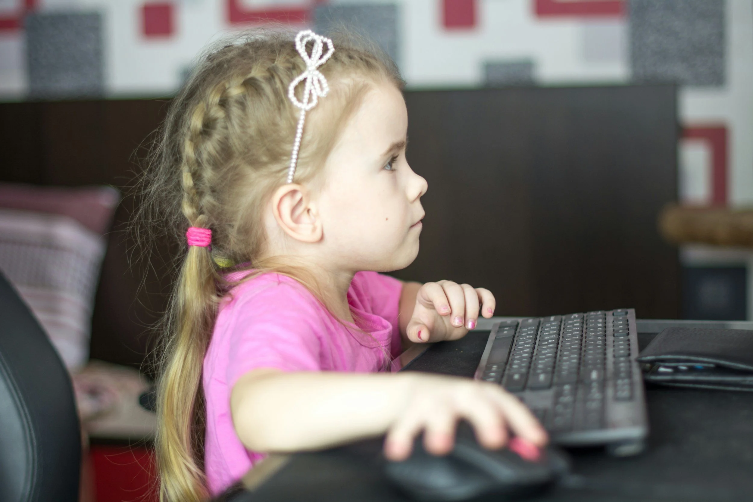 A young girl with braided blonde hair, wearing a pink shirt, sitting at a desk and using a computer keyboard and mouse.