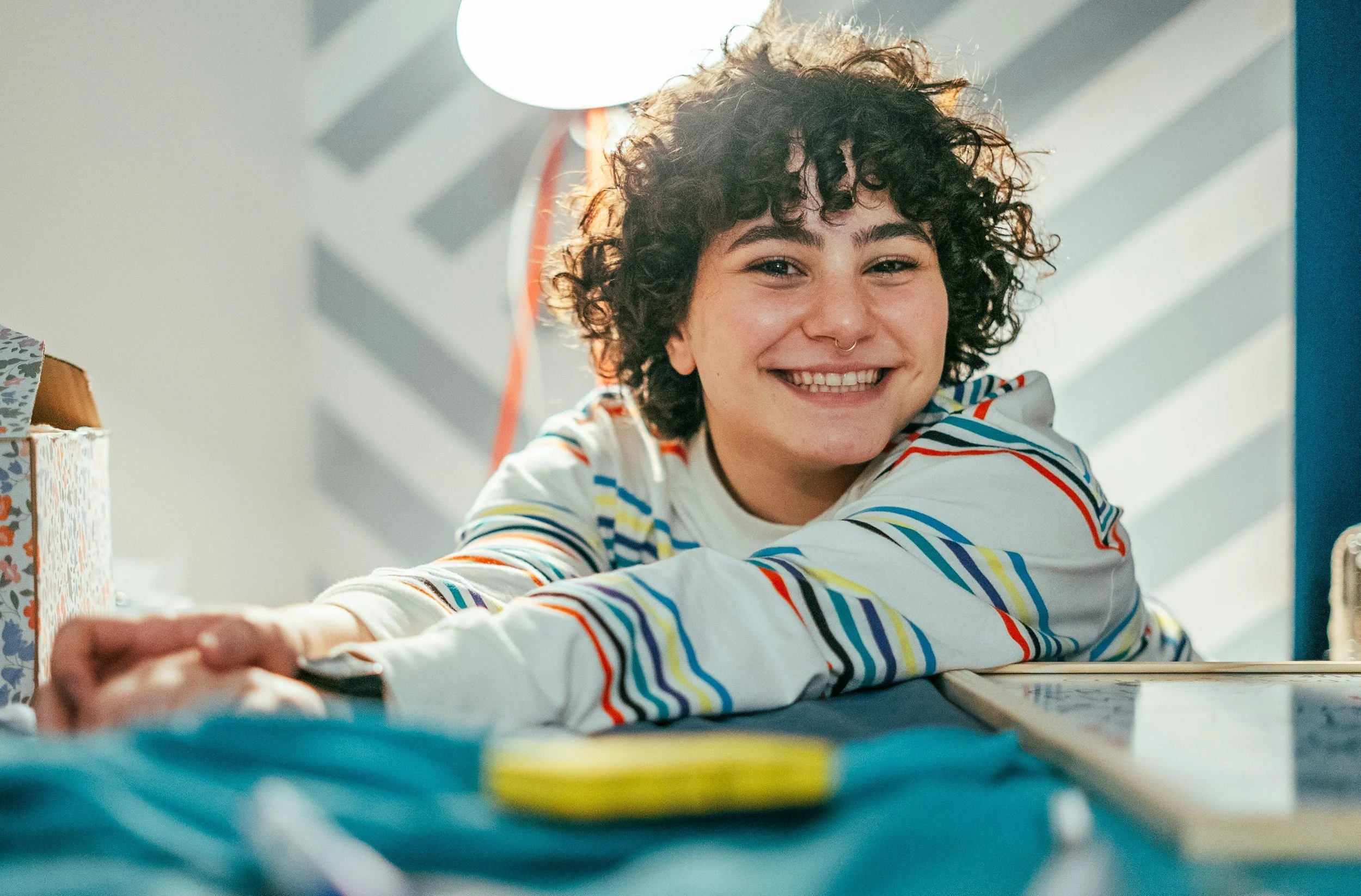 Smiling young woman with curly hair and nose ring, reaching forward on a table, with a bright background and a lamp behind her.