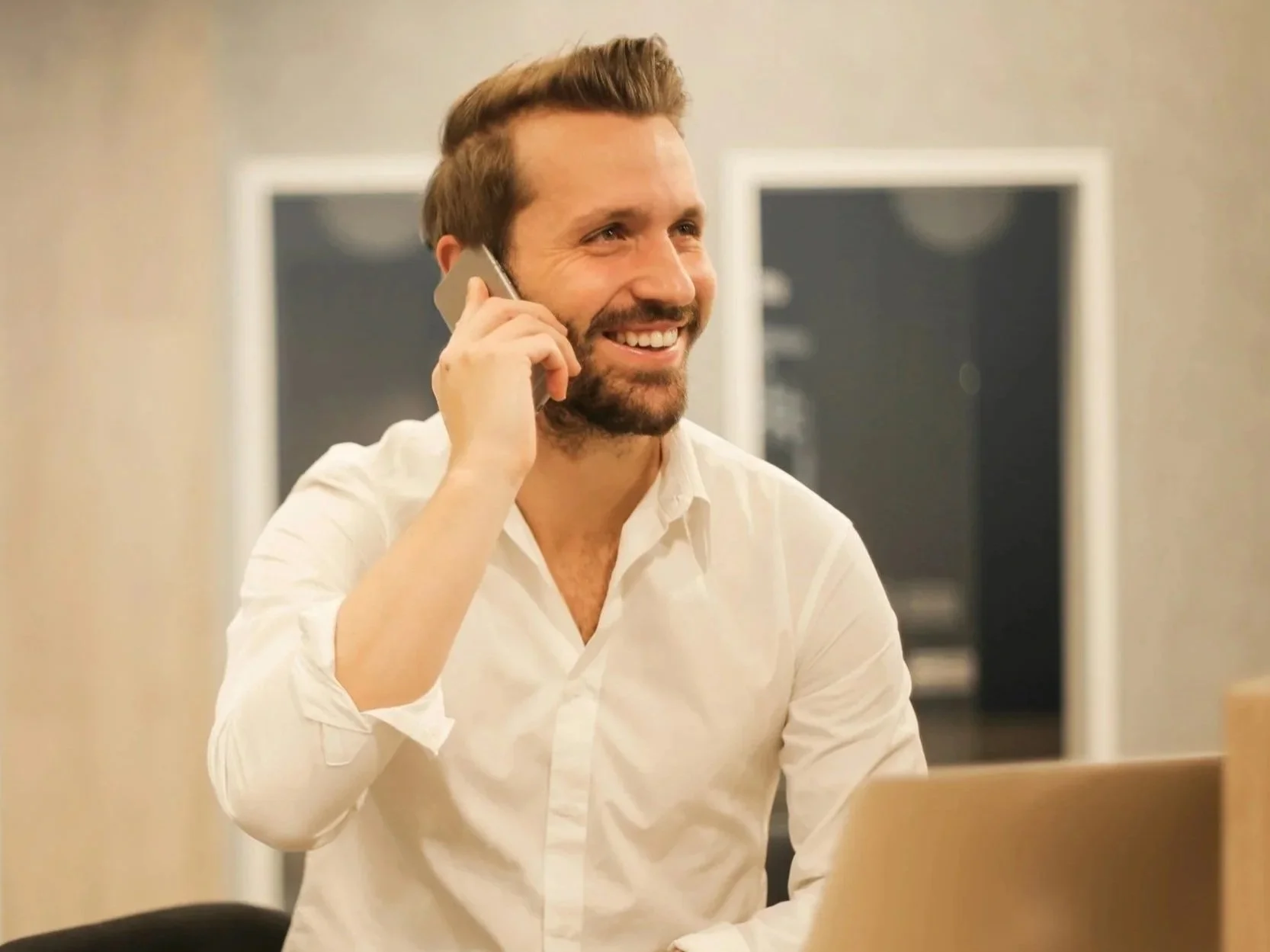 A smiling man with a beard and dressed in a white shirt talking on a landline phone in an office.