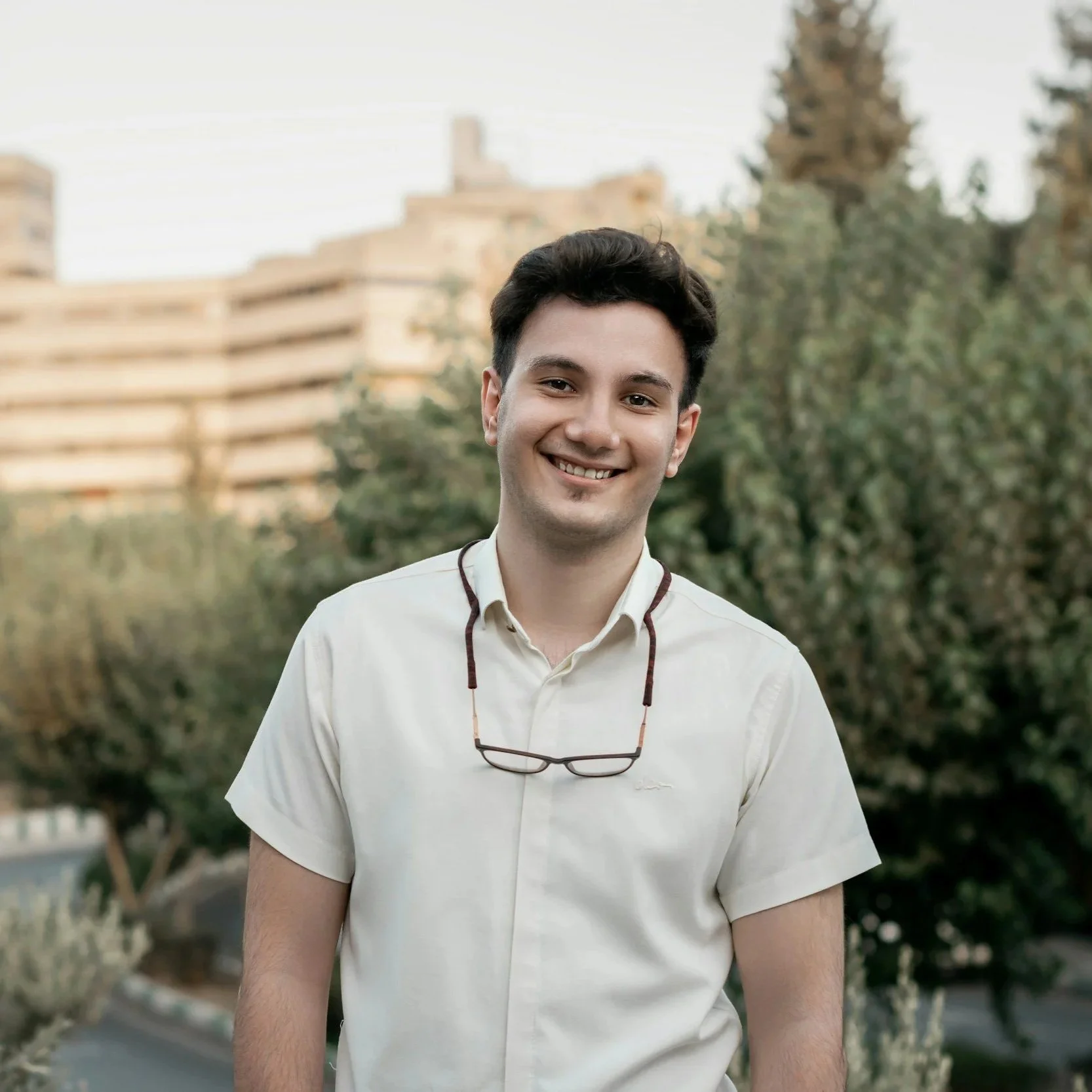Young man smiling outdoors with glasses around his neck, trees and buildings in the background.