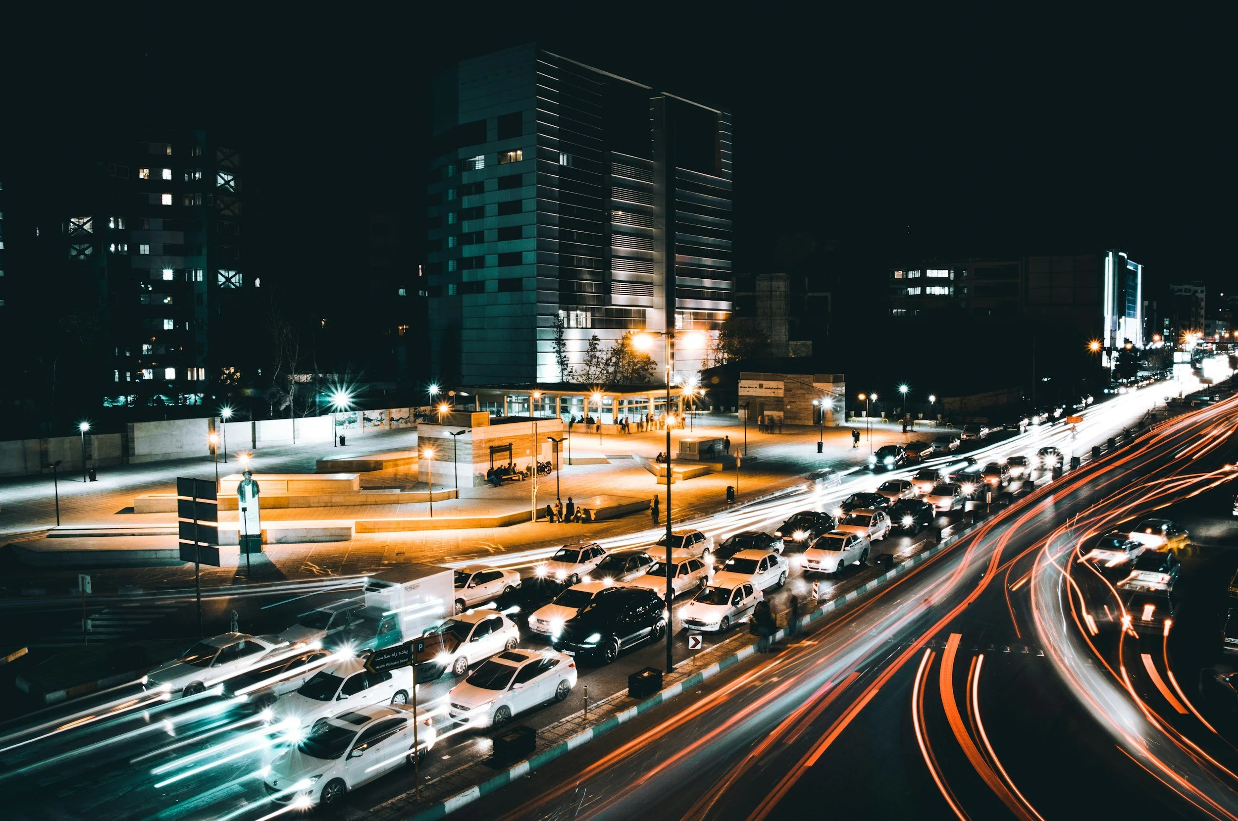 Night time city scene with a busy street filled with moving cars, light trails from long exposure, and modern high-rise buildings in the background.