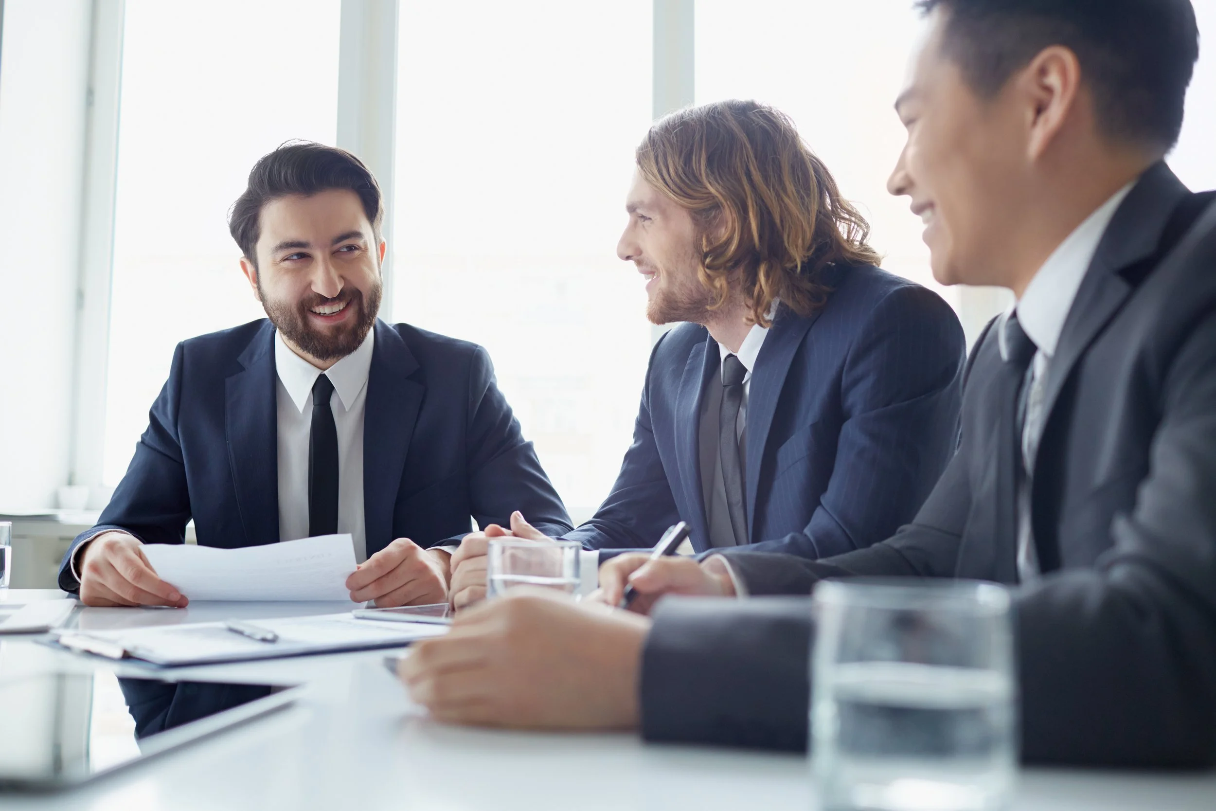 Three businessmen in suits smiling and talking during a meeting in a bright office conference room.