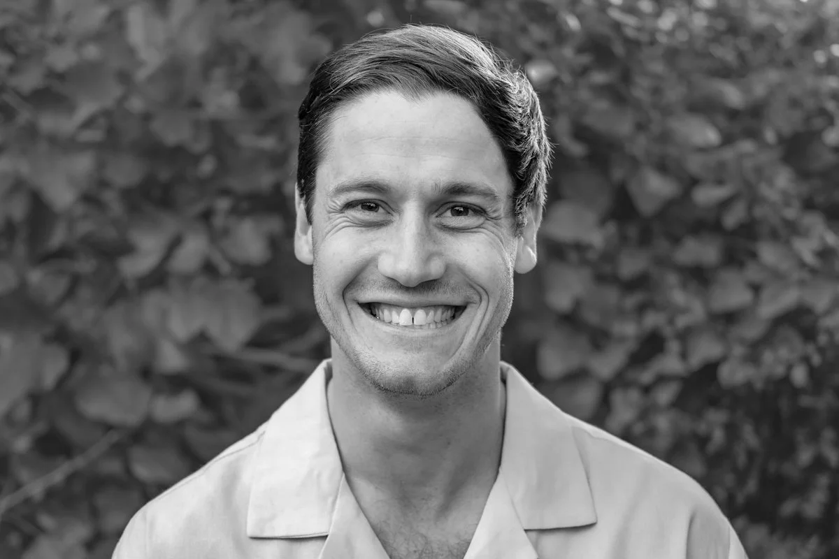 Black and white photo of smiling man with short hair, wearing a collared shirt, standing outdoors with blurred foliage background.