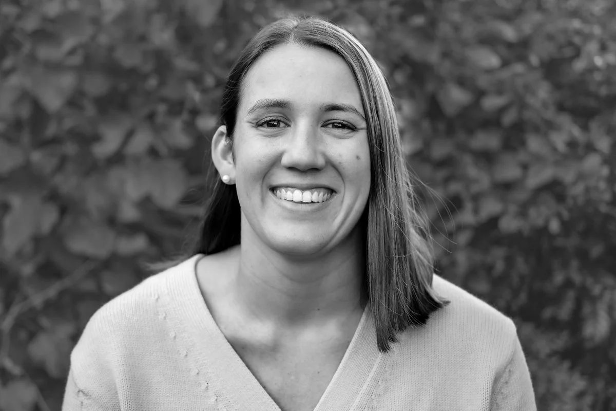 Black and white portrait of a smiling woman with shoulder-length hair, wearing a light-colored top and pearl earrings, against a blurred foliage background.