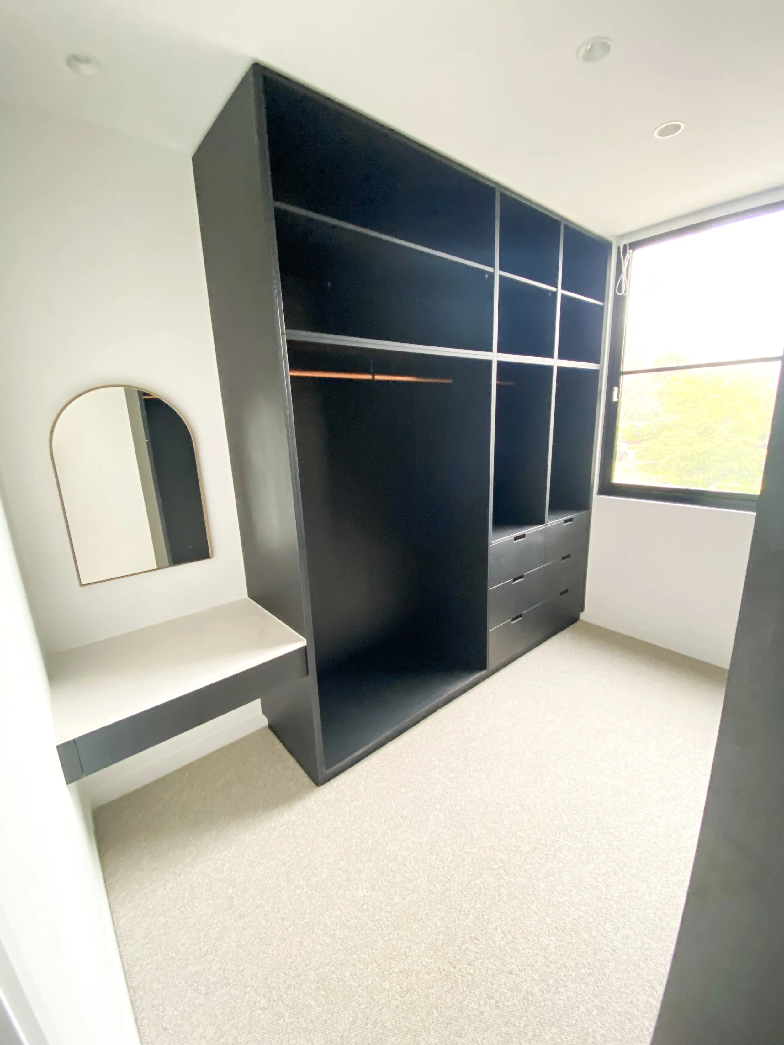 Empty black shelving unit with drawers below, next to a window, in a room with beige carpet and white walls.