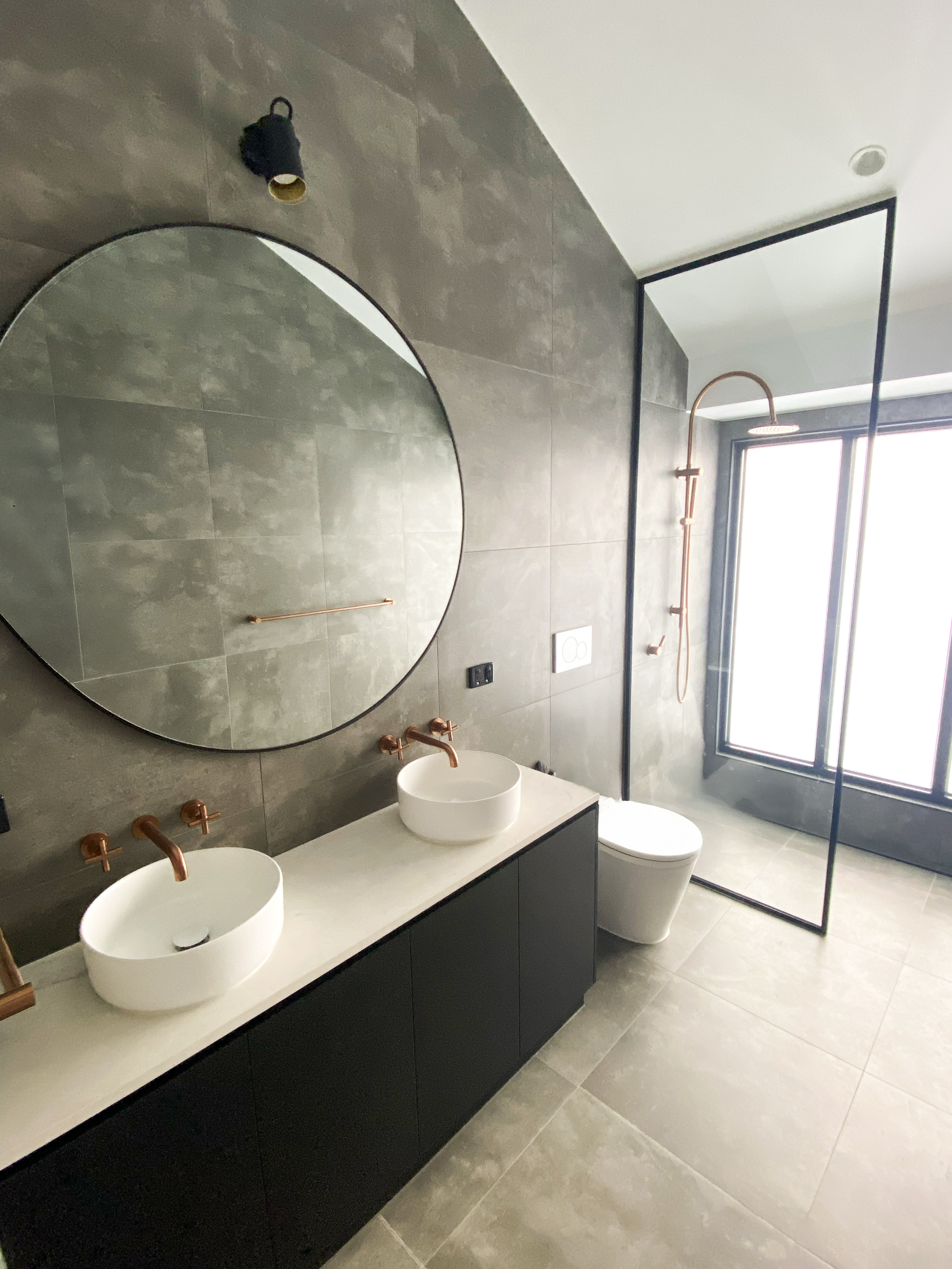 Modern bathroom with gray tiled walls and floor, a black vanity with two white vessel sinks, a round mirror, a toilet, a large window, a black metal frame dividing the shower area, a wall-mounted showerhead, and a dark light fixture.