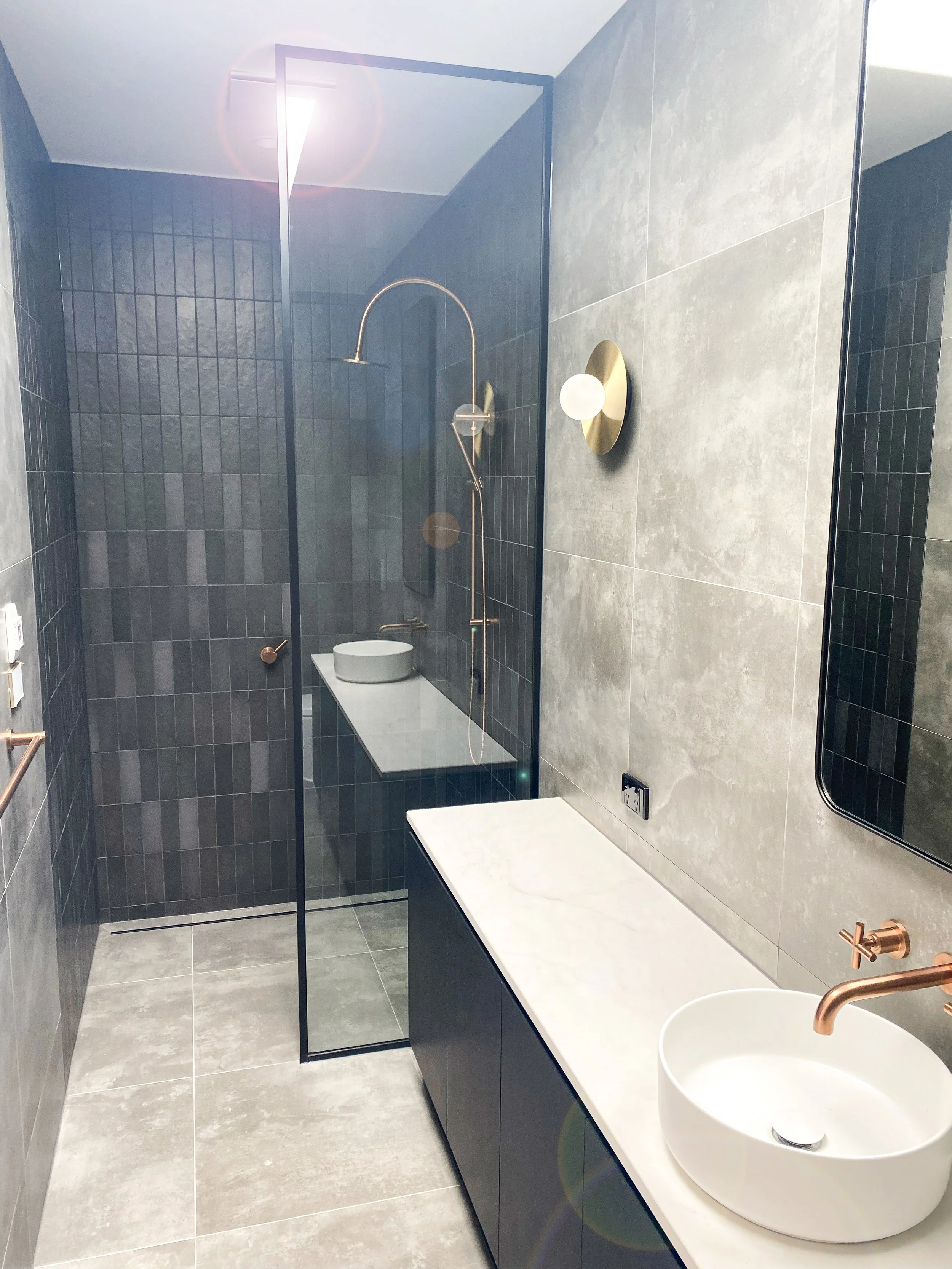 Modern bathroom with grey tiles on the walls and floor, a black-framed shower enclosure with a copper shower fixture, a white countertop with a round vessel sink, and a gold wall-mounted light fixture.