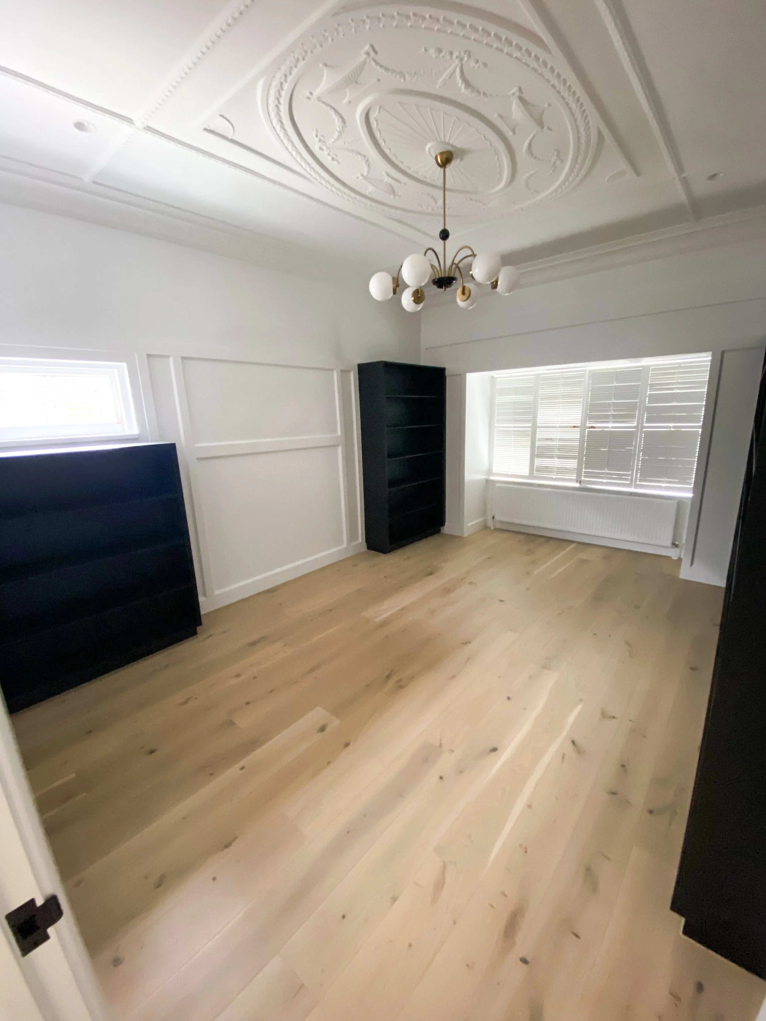 Empty room with wooden floor, black shelving unit, window with blinds, ceiling light fixture, and decorative ceiling design.