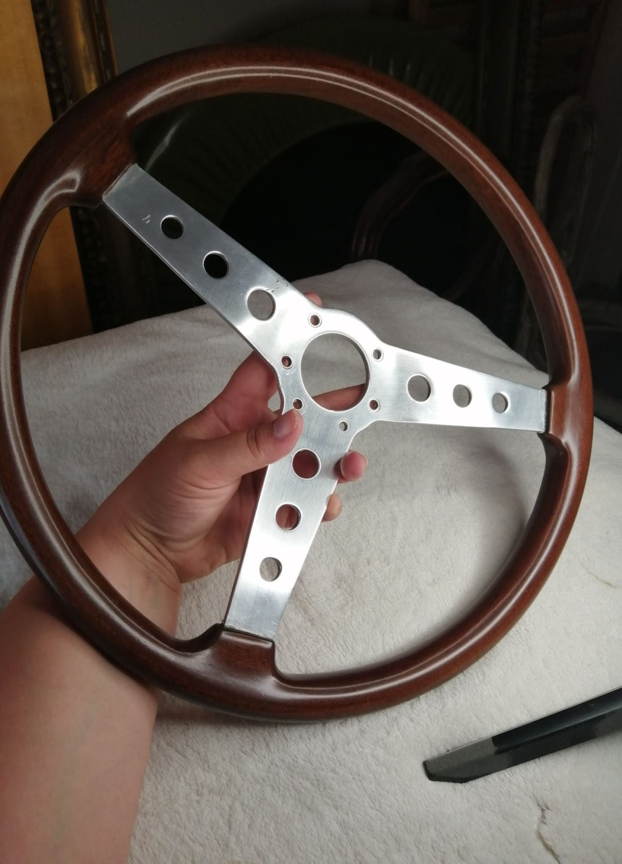 A hand holding a vintage wooden steering wheel with a metal center. The wheel has a dark polished wood rim and the metal center has multiple holes for attaching to a vehicle's steering column.