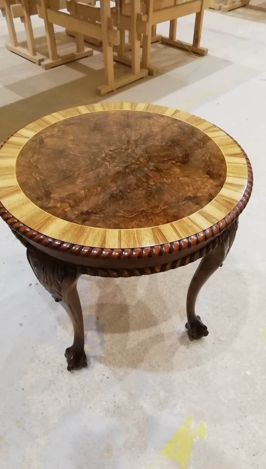 A round wooden table with carved legs and a decorative rim, featuring a dark wood center and a lighter wood border, set on a concrete floor with a background of stacked wood furniture.